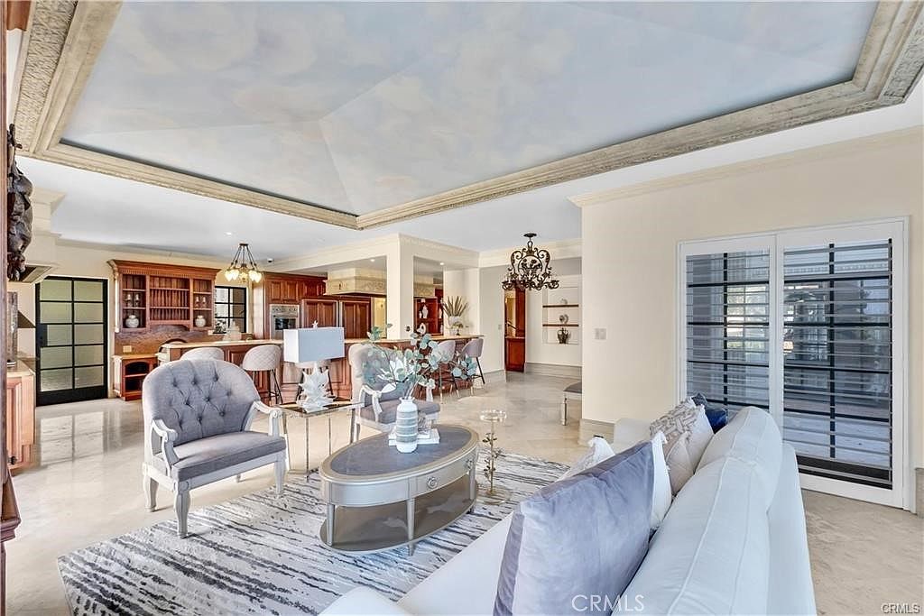 This spacious, open-concept living area features a dramatic tray ceiling painted with a cloud mural and framed by decorative molding. The room is furnished with a plush white sofa, a grey tufted armchair, and a central oval coffee table, all set atop a textured area rug. Large sliding glass doors provide access to the outdoors, while the background reveals a well-appointed kitchen with wooden cabinetry and a breakfast bar.