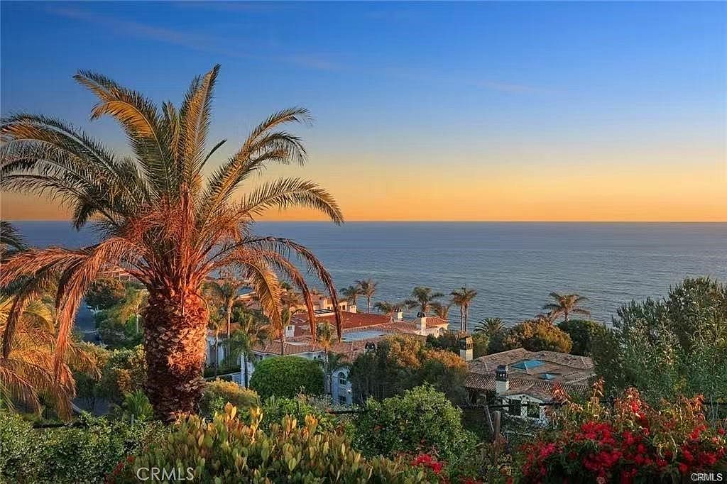 This elevated, wide-angle shot captures a stunning coastal sunset view overlooking a Mediterranean-style estate. In the foreground, a prominent palm tree and vibrant red bougainvillea frame the scene, while the mid-ground reveals the tiled roofs of the property nestled among lush greenery. The horizon line where the calm ocean meets the soft, golden-hued sky creates a serene and luxurious atmosphere.