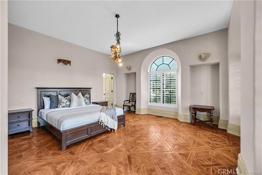 This spacious primary bedroom features intricate, patterned wood flooring and light-toned walls, creating an elegant and airy atmosphere. A large bed with a dark wood frame serves as the focal point, complemented by a unique, ornate chandelier hanging from the ceiling. The room includes a large arched window with shutters and a recessed alcove, offering a sophisticated architectural aesthetic.