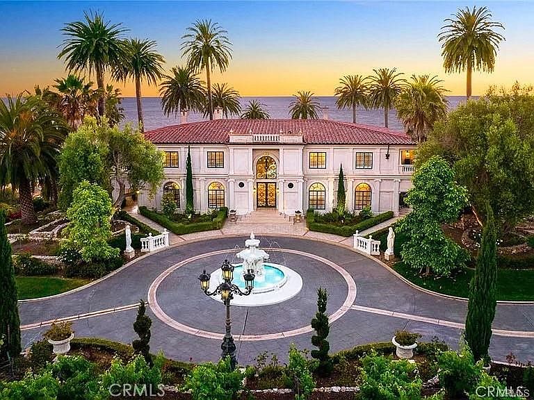 This grand Mediterranean-style estate features a symmetrical facade with a terracotta tile roof and a prominent arched entryway. A circular driveway centers around an ornate tiered fountain, framed by manicured hedges and mature palm trees. The property offers a stunning oceanfront perspective, with the vast sea visible in the background under a warm sunset sky.