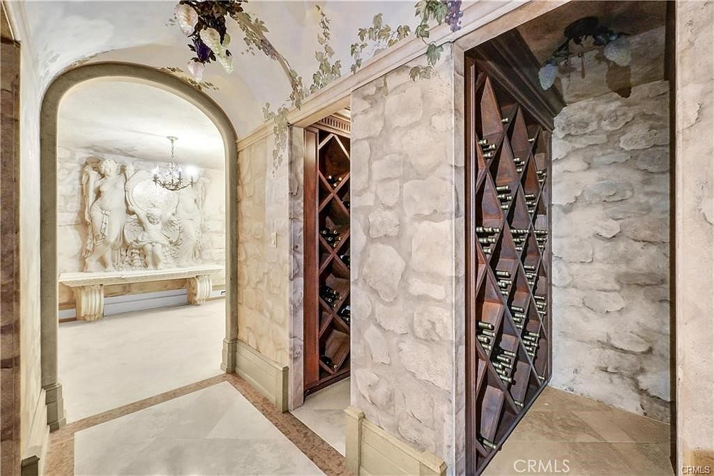 This sophisticated wine cellar features a dramatic arched entryway leading to a secondary room adorned with a classical-style stone relief sculpture. The space is characterized by rustic stone walls, custom diamond-patterned wooden wine racks, and decorative painted vines trailing across the ceiling. The perspective captures the transition between the wine storage area and the adjacent gallery, creating an atmosphere of old-world elegance.