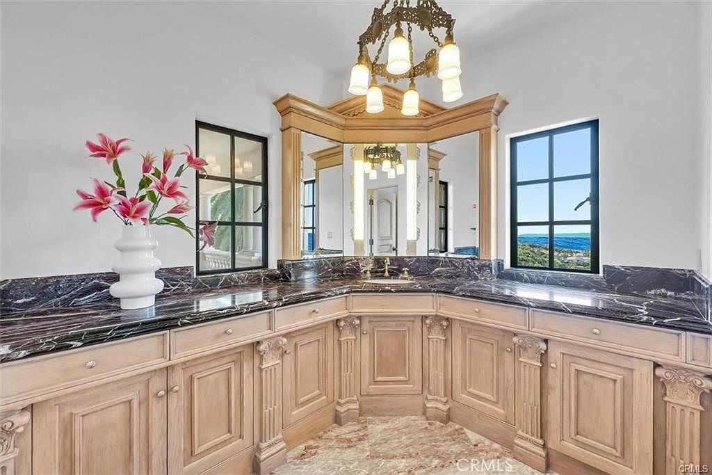 This elegant bathroom vanity area features light wood cabinetry with ornate detailing and a dark, veined stone countertop. A multi-faceted mirror arrangement is framed by wood trim, complemented by a decorative chandelier and large windows that provide natural light and scenic views. The space is accented by a white vase with pink lilies, creating a sophisticated and refined atmosphere.