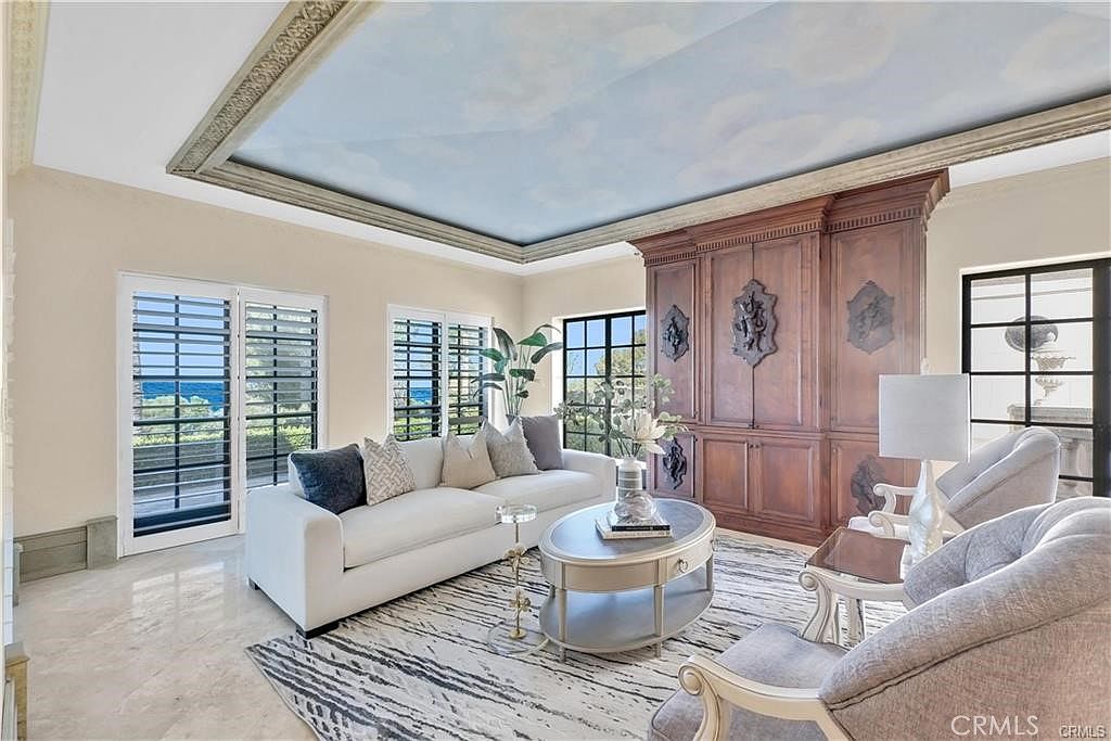 This elegant living room features a large white sofa, a central oval coffee table, and two armchairs, all arranged on a patterned area rug. A prominent, dark wood built-in cabinet unit serves as a focal point, while the ceiling is uniquely decorated with a painted sky mural framed by decorative molding. Large windows with shutters allow for natural light and offer views of the outdoors, creating a bright and sophisticated atmosphere.