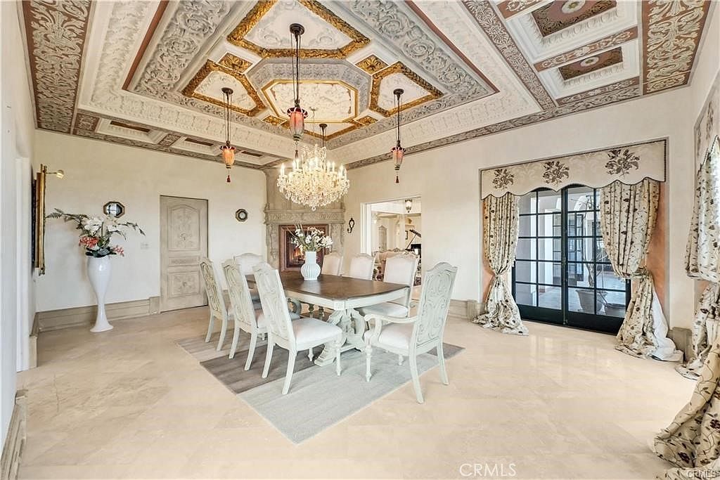 This elegant dining room features a formal table set for six, positioned beneath an ornate crystal chandelier and a highly detailed, coffered ceiling with gold-leaf accents. The space is anchored by a stone fireplace and large glass doors draped in patterned curtains that allow natural light to fill the room. The neutral stone flooring and sophisticated architectural molding create a luxurious and refined atmosphere suitable for formal entertaining.