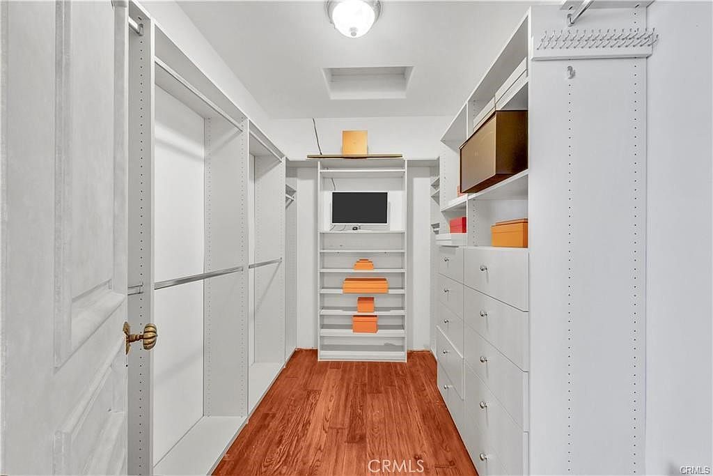 This walk-in closet features custom white shelving and cabinetry, providing ample storage space with hanging rods and multiple drawers. The room is finished with warm-toned wood flooring and a central ceiling light fixture. A small television is mounted on the back wall, surrounded by open shelving that displays several orange storage boxes.