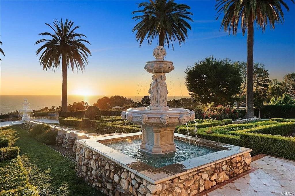 This expansive garden features a prominent tiered stone fountain as its centerpiece, set within a manicured landscape of structured hedges and lush green lawns. Tall palm trees frame the scene, which overlooks a serene ocean horizon during a vibrant sunset. The stone-walled fountain basin and elegant statuary create a sophisticated, resort-like atmosphere perfect for outdoor relaxation.
