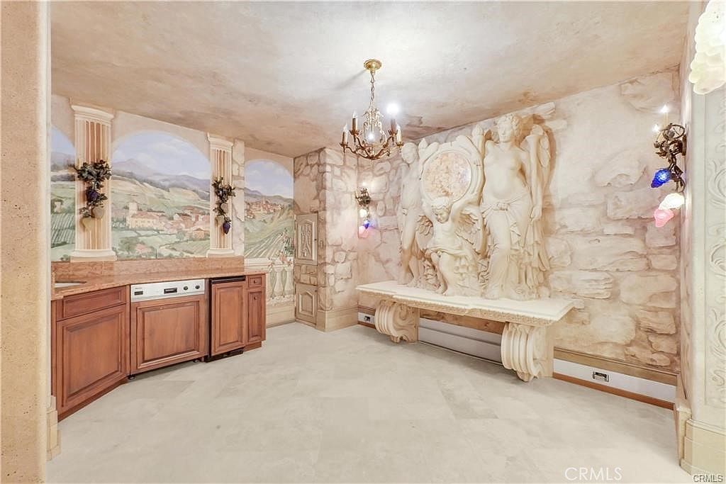 This unique interior space features a highly decorative, Mediterranean-inspired aesthetic with a large, ornate stone relief sculpture serving as a focal point above a stone console table. The room includes a wet bar area with wood cabinetry and a mural depicting a rolling landscape, all set against textured stone walls and a soft, neutral-toned floor. A classic chandelier hangs from the ceiling, contributing to the dramatic, old-world atmosphere of the room.