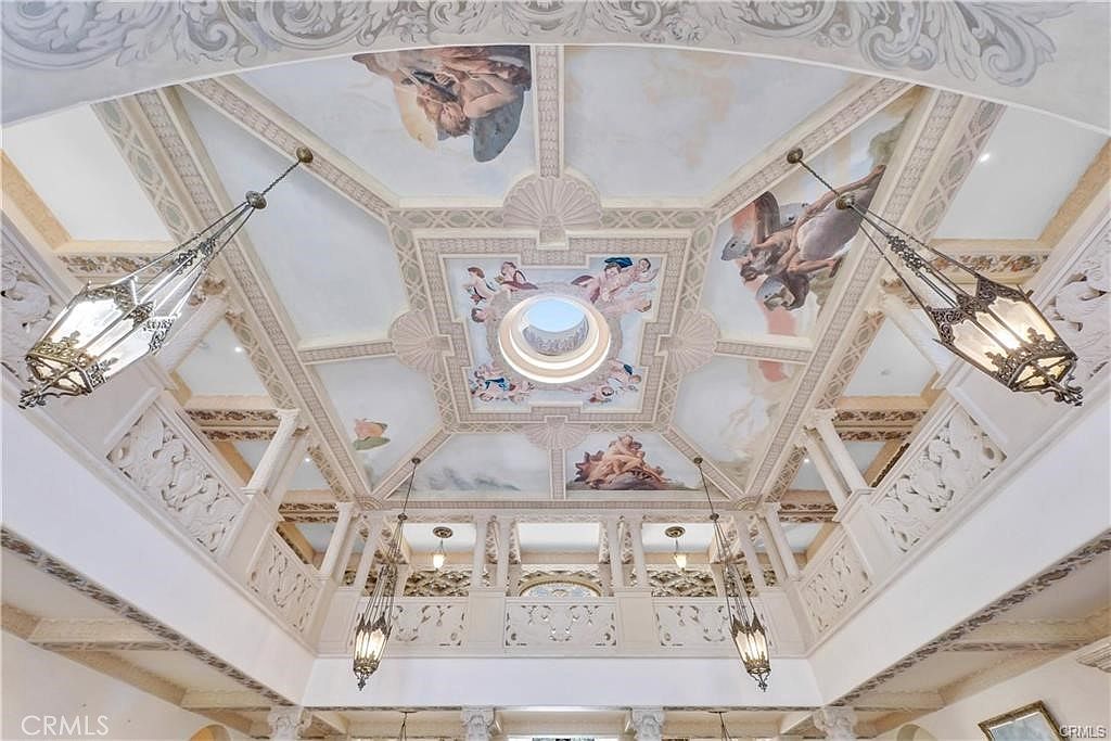 This image captures an ornate, multi-tiered ceiling featuring elaborate hand-painted frescoes depicting classical figures and cherubs. The architectural design includes a central circular skylight, intricate molding, and decorative corbels, all illuminated by several hanging lantern-style chandeliers. The perspective is looking directly upward, emphasizing the grand scale and opulent, classical aesthetic of the interior space.