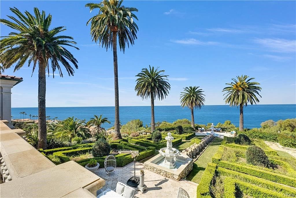 This elevated view captures a meticulously landscaped formal garden featuring geometric boxwood hedges and a central stone fountain. Tall palm trees frame the expansive, unobstructed view of the deep blue ocean horizon. The scene includes a stone-paved patio area in the foreground, creating a luxurious and serene outdoor living space.