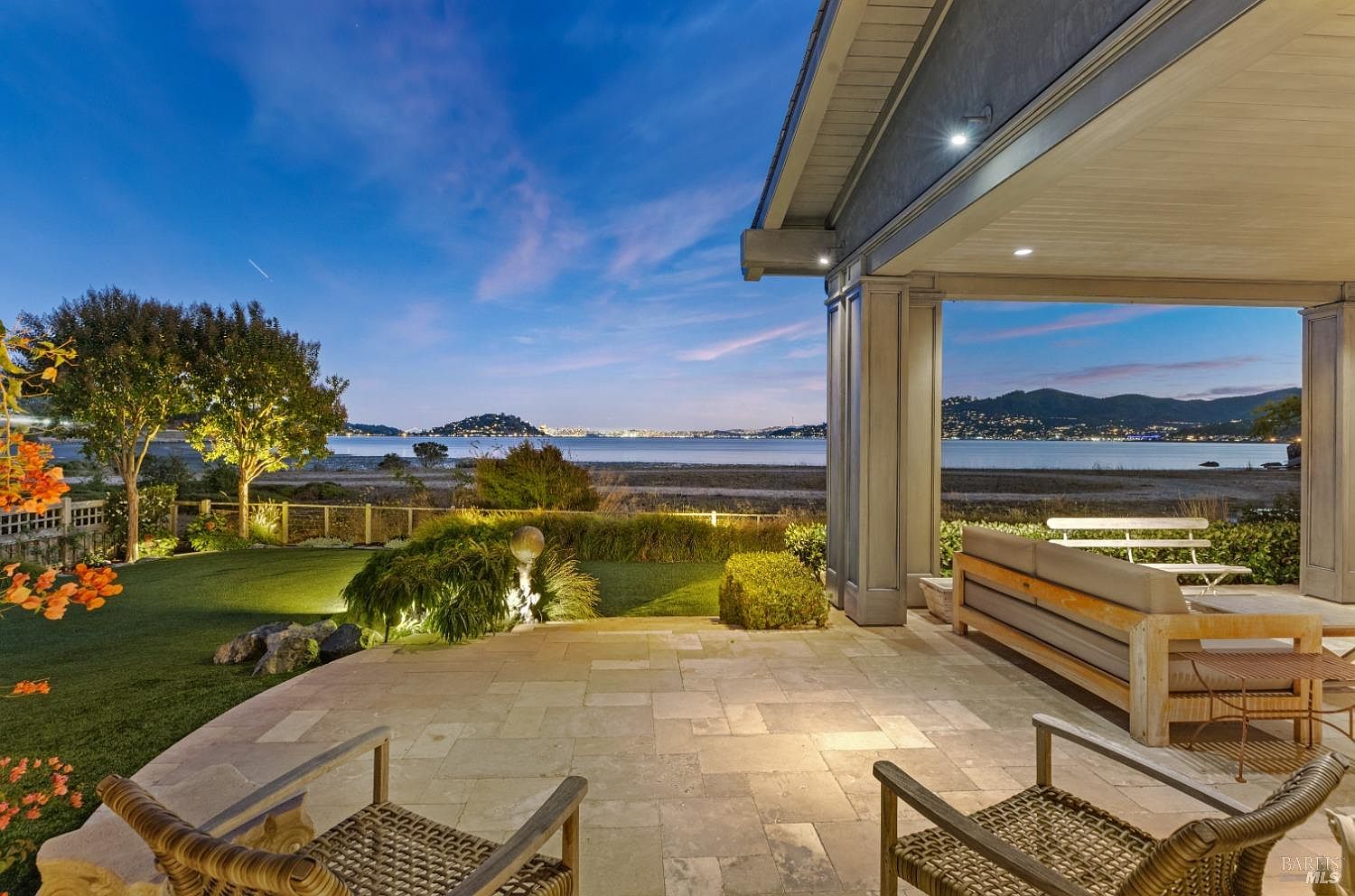 This image showcases a luxurious outdoor patio area at dusk, featuring stone flooring and comfortable wicker furniture. The patio offers stunning waterfront views with city lights visible in the distance, creating a serene and upscale ambiance. The well-manicured lawn and garden add to the property's appeal, making it an ideal space for relaxation and entertainment.