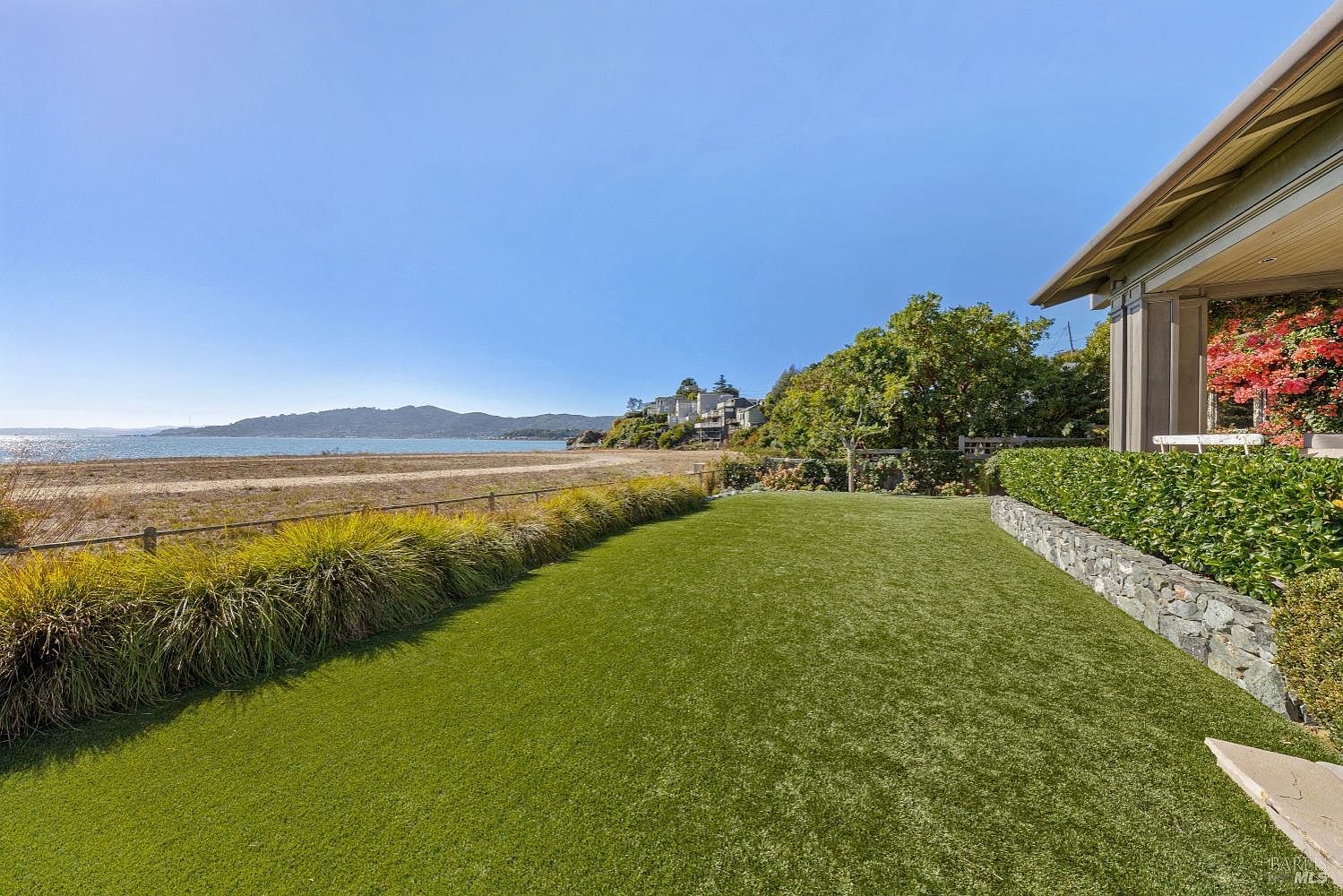 This image showcases a well-maintained yard with lush green grass extending towards a sandy beach and ocean view. A neatly trimmed hedge and a stone retaining wall add structure to the landscape, while a portion of the house is visible on the right, suggesting a private and luxurious setting. The scene evokes a sense of tranquility and upscale coastal living.