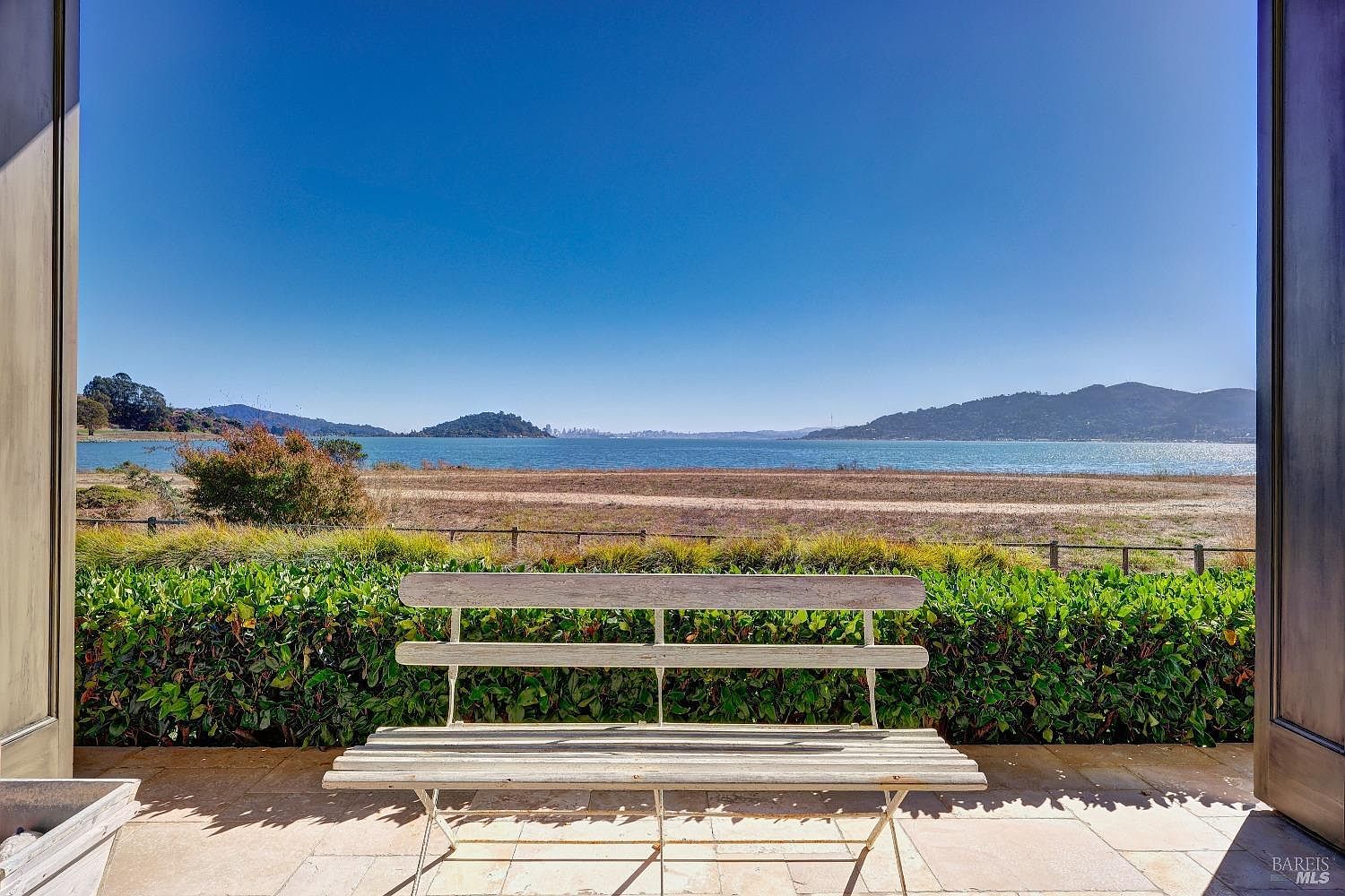 This image showcases a serene patio or balcony with a stunning waterfront view. A weathered wooden bench sits in the foreground, inviting relaxation and contemplation. The scene is framed by open doors, adding a sense of openness and connection to the outdoors, while lush greenery provides a natural border, enhancing the tranquil atmosphere.