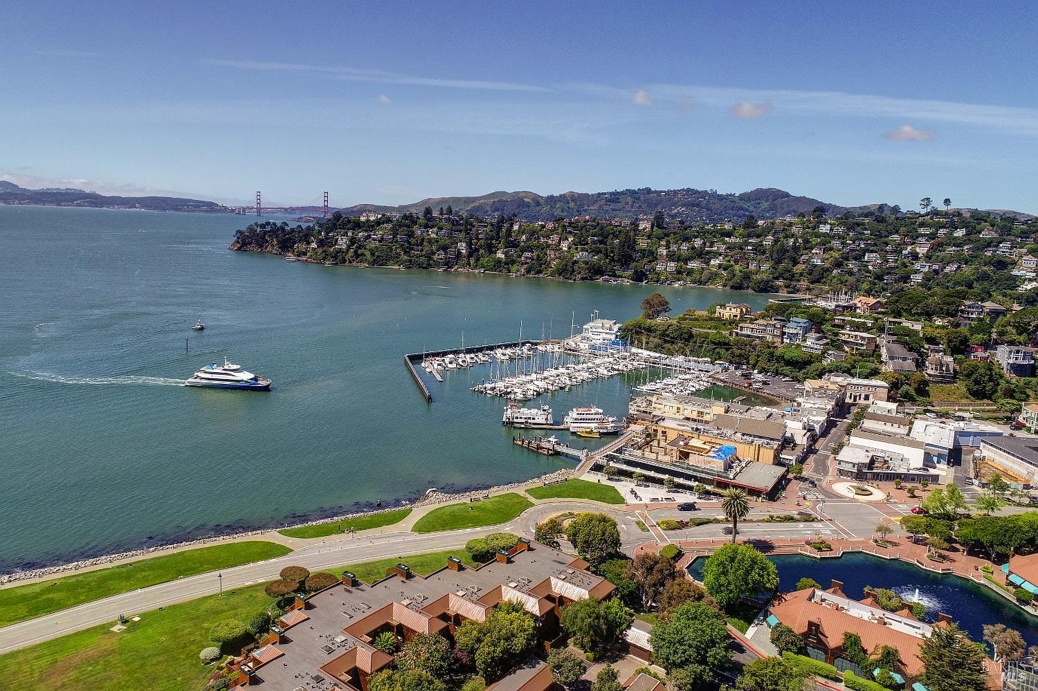 This aerial shot showcases a picturesque coastal town with a marina filled with boats, surrounded by lush green hills and residential areas. A ferry is seen sailing on the water, and the Golden Gate Bridge is visible in the distance. The image provides a comprehensive view of the area's natural beauty and urban development, highlighting its appeal as a desirable location.