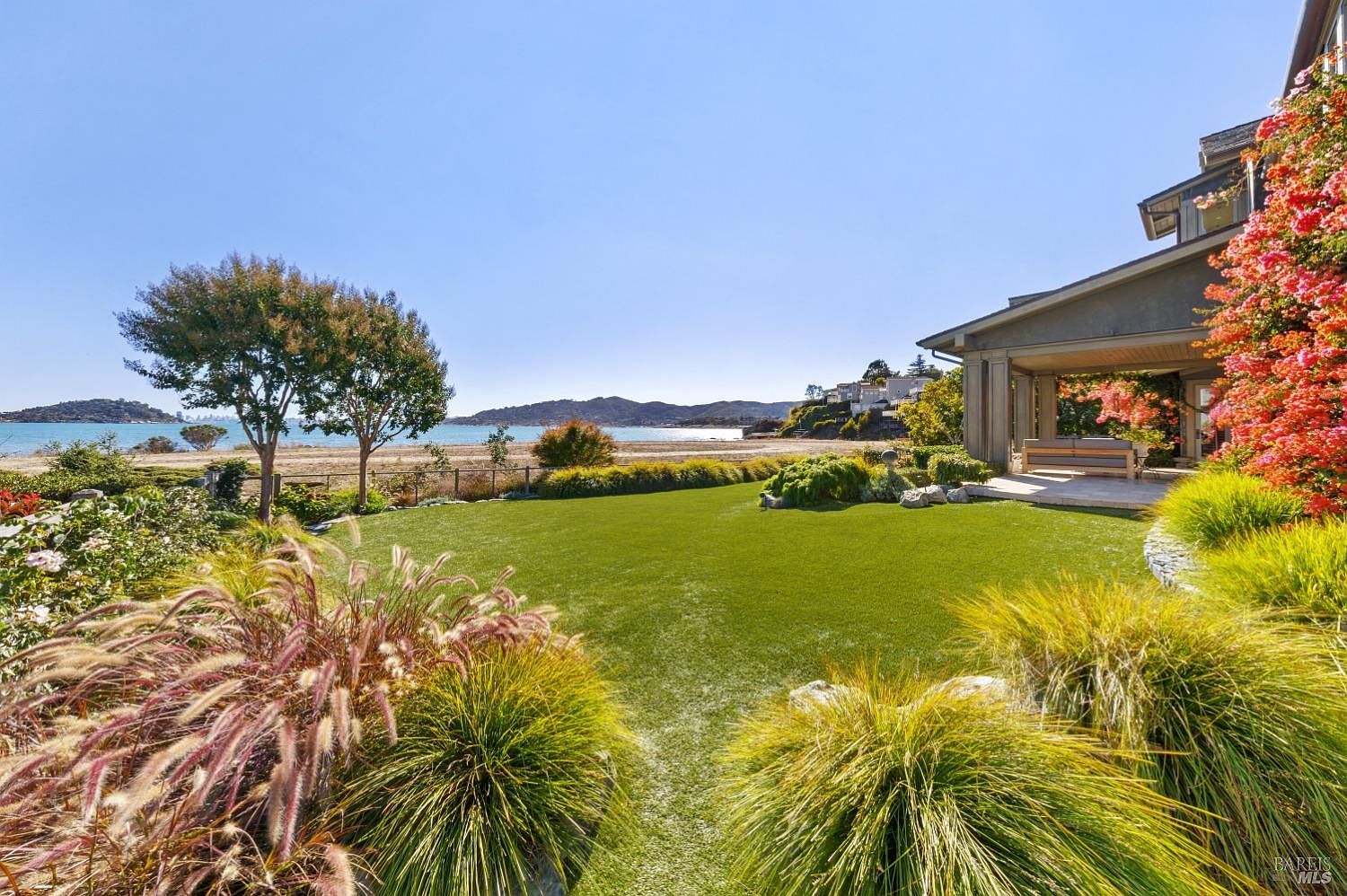 This is a beautiful exterior shot of a well-manicured yard and garden leading to a beach. The lush green lawn is framed by ornamental grasses and a mature tree, with a glimpse of the ocean and coastline in the background. A portion of the house is visible on the right, adorned with vibrant flowering vines, suggesting a luxurious and serene coastal living experience.