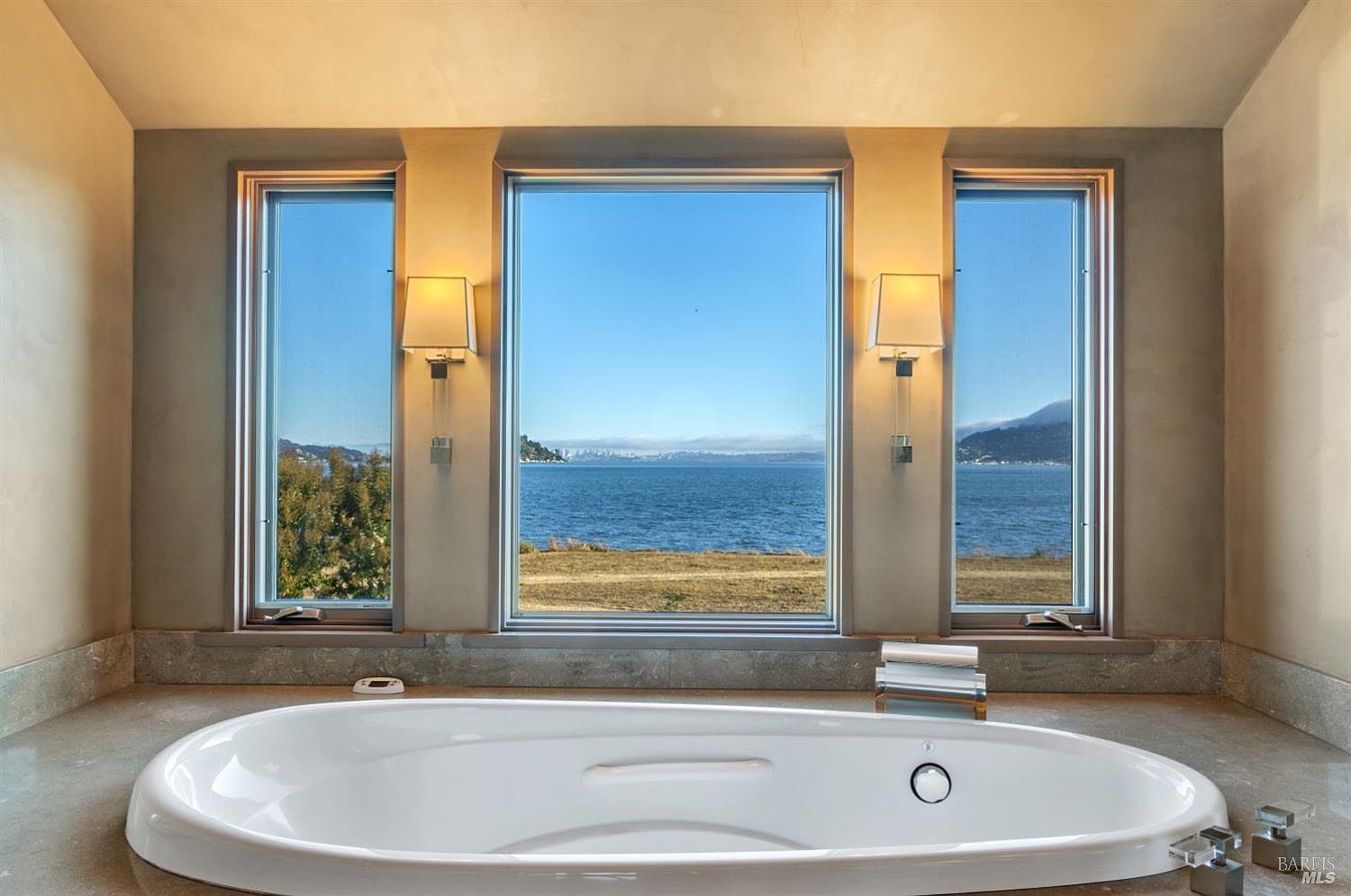 This is a luxurious primary bathroom featuring a large, oval-shaped soaking tub positioned in front of three expansive windows offering stunning views of the ocean. The room is decorated in neutral tones, creating a serene and spa-like atmosphere. The perspective is from the entrance of the bathroom, showcasing the tub as the focal point.