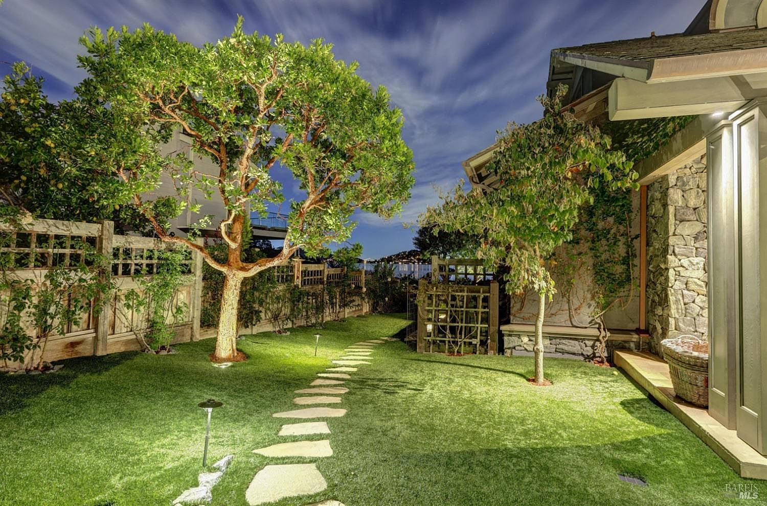 This is a beautifully landscaped backyard at dusk, featuring a stone pathway leading through a lush green lawn. Mature trees are strategically placed, with subtle lighting enhancing their form. A wooden fence and a stone-clad portion of the house add texture and character to the scene, creating a serene and inviting outdoor space.