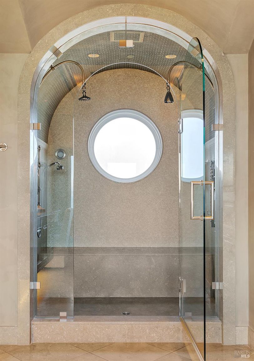 This is a luxurious primary bathroom featuring a walk-in shower with a unique arched design and a round window. The shower is enclosed with clear glass panels and has dual shower heads. The walls and ceiling are covered in a textured, neutral-toned material, creating a spa-like atmosphere.