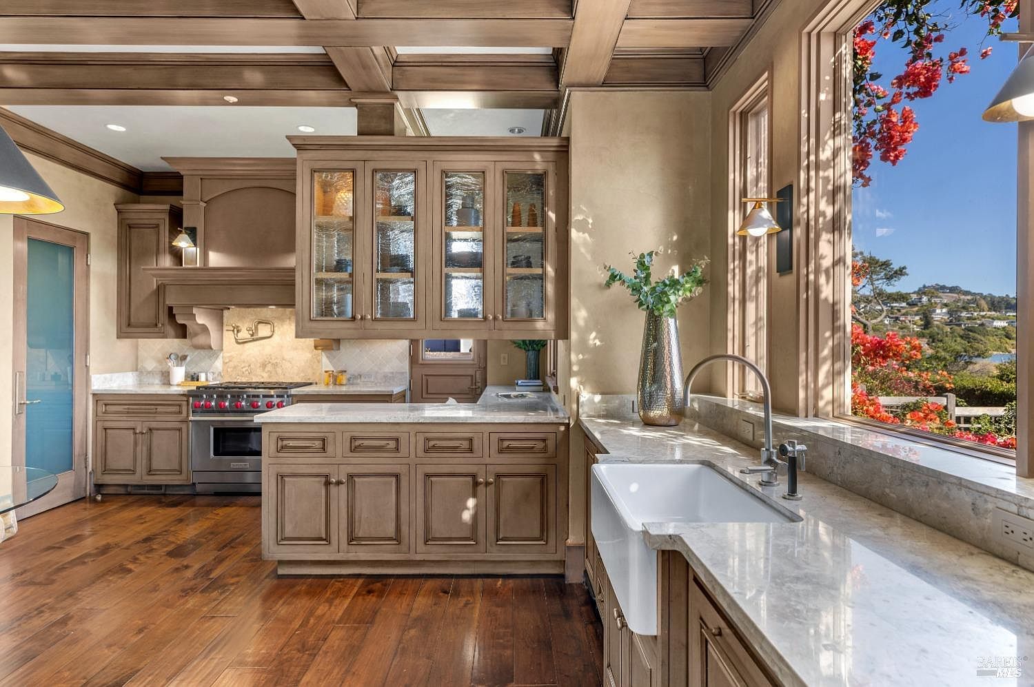 This is a well-appointed kitchen featuring custom cabinetry with glass-fronted upper cabinets, marble countertops, and a farmhouse sink. The kitchen is bathed in natural light from a large window overlooking a scenic view, and the hardwood floors add warmth to the space. The overall impression is one of luxury and functionality.