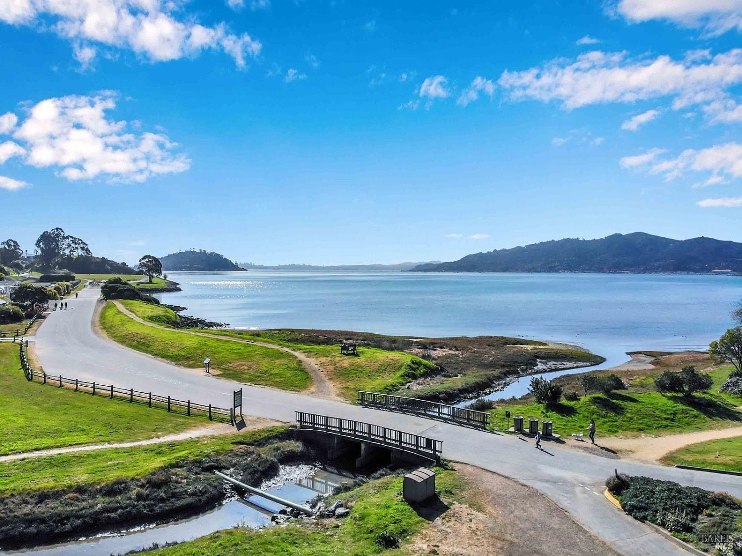 This aerial view showcases a scenic coastal landscape with a winding road leading to a bridge over a small waterway. Lush green grass and trees line the road, contrasting with the blue water and distant hills. The image captures a serene and picturesque environment, ideal for highlighting the natural beauty surrounding a property.