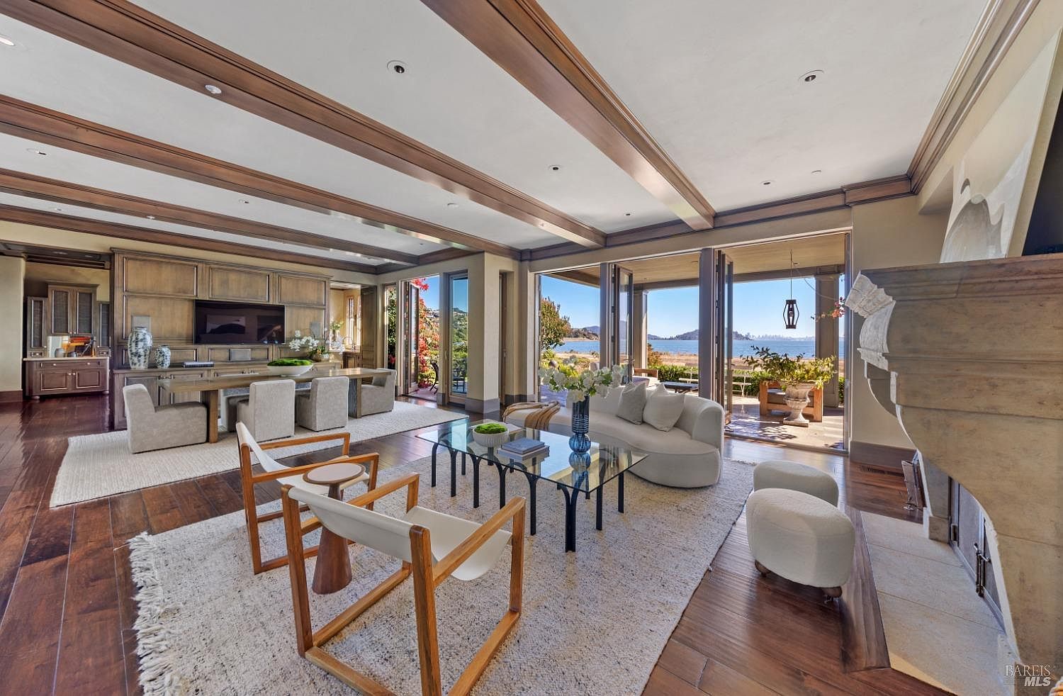 This is a spacious living room featuring hardwood floors, exposed beam ceilings, and a large stone fireplace. The room is furnished with a curved sofa, glass coffee table, and modern chairs. Large folding doors open to an outdoor patio with ocean views, creating a seamless indoor-outdoor living experience.