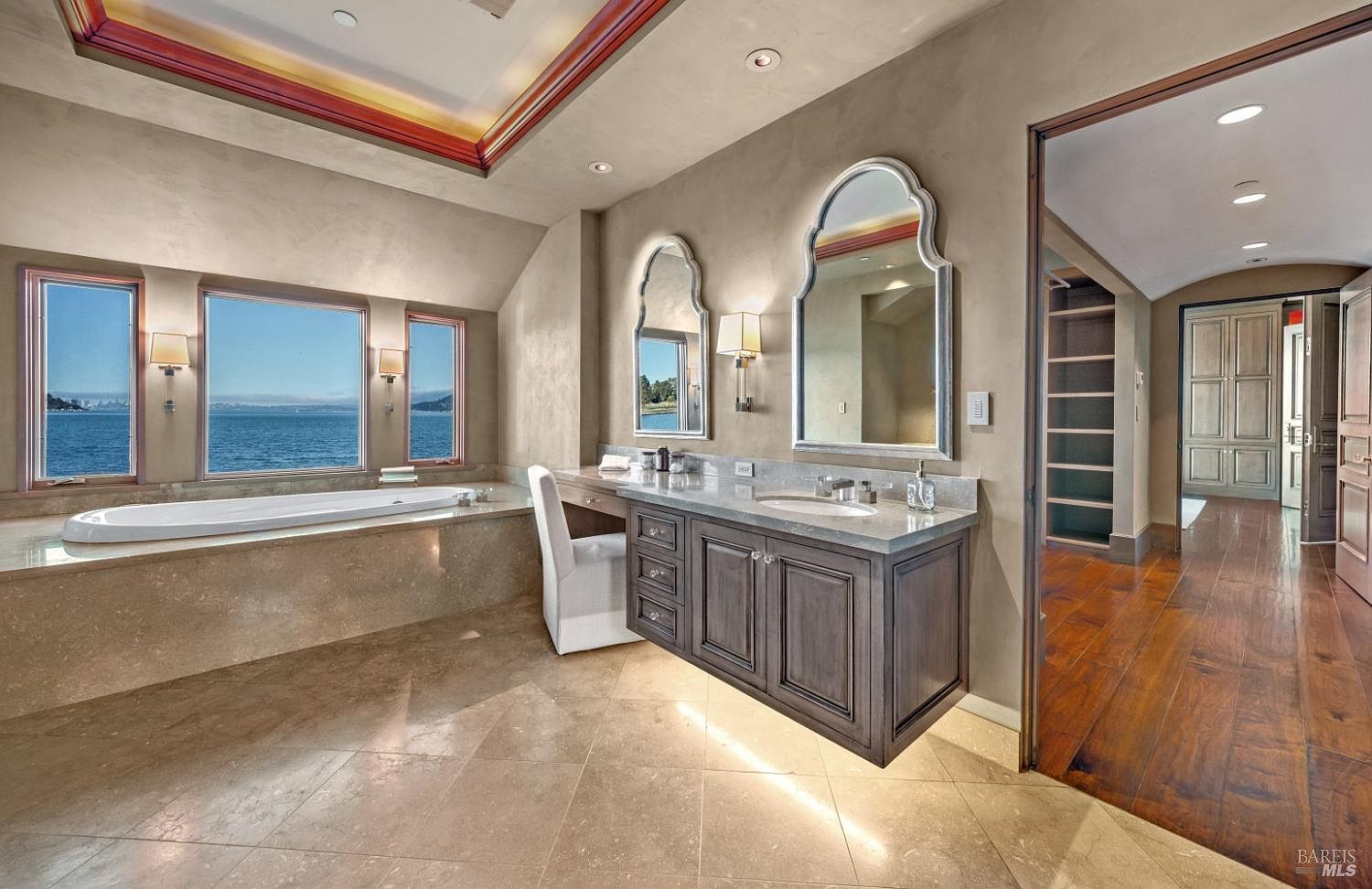 This is a luxurious primary bathroom featuring a large soaking tub positioned beneath three windows offering a scenic water view. The bathroom includes a vanity with dual sinks, custom cabinetry, and decorative mirrors. The flooring is tiled, and the overall design exudes elegance and tranquility.