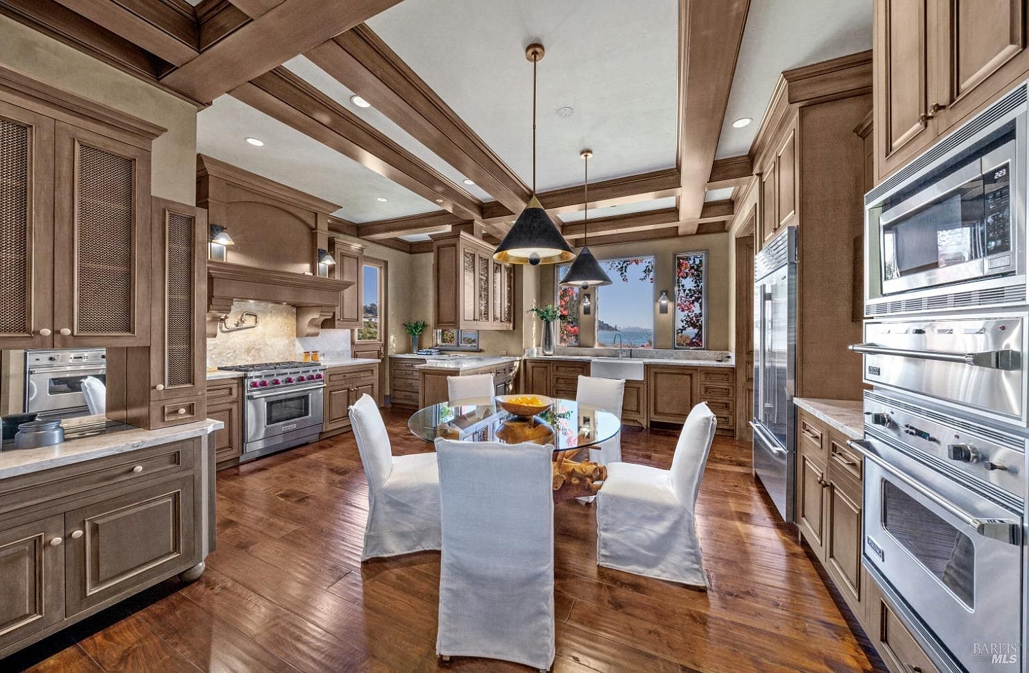 This is a well-appointed kitchen featuring custom wood cabinetry, stainless steel appliances, and a central dining area with a glass-top table and white upholstered chairs. The ceiling has exposed wooden beams with recessed lighting, and pendant lights hang above the table. The kitchen offers a blend of traditional and modern design elements, creating a warm and inviting space.