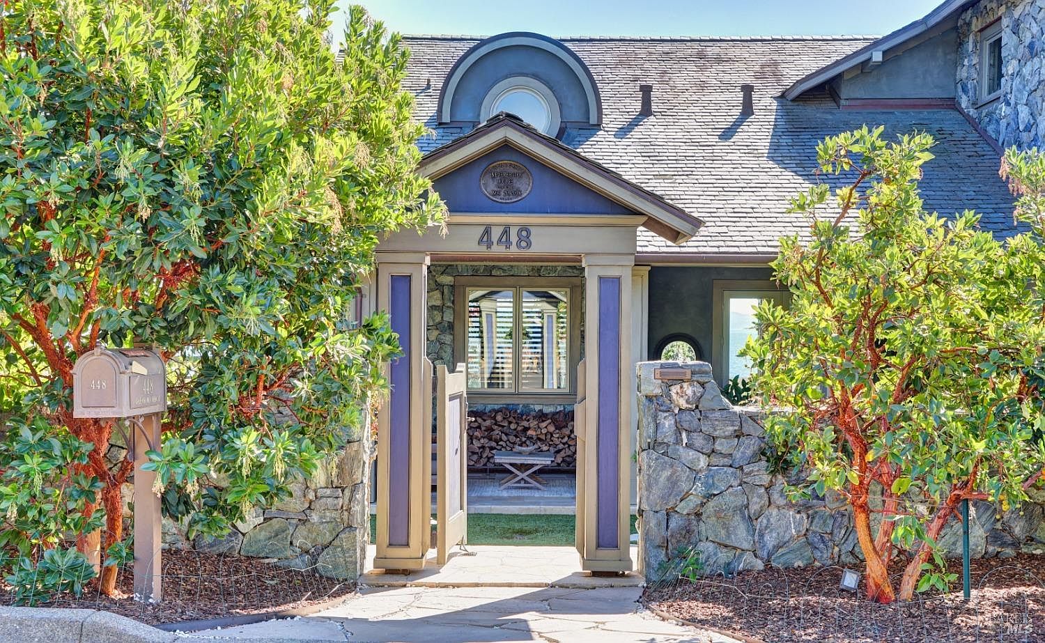The image showcases the entryway of a charming home, featuring a stone archway with wooden gates and the house number '448' prominently displayed. Lush greenery flanks the entrance, adding to the property's curb appeal. The overall impression is inviting and picturesque, suggesting a well-maintained and cared-for residence.
