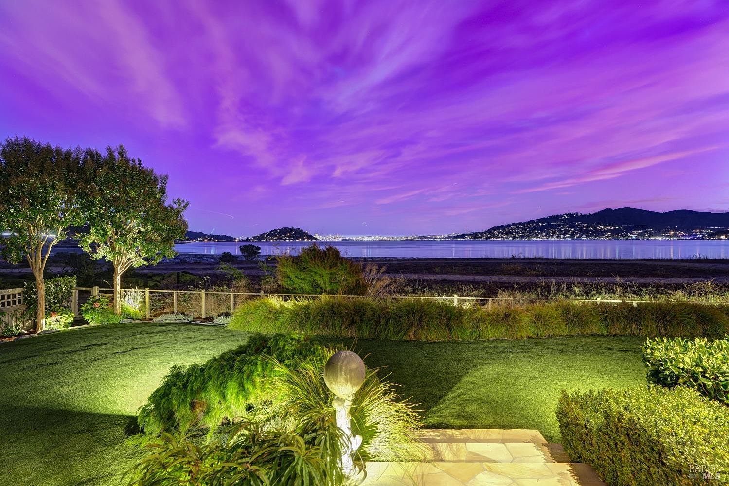 This image showcases a beautifully landscaped backyard with a lush green lawn, manicured bushes, and a decorative stone pathway. A wooden fence borders the property, offering a view of the water and distant city lights under a vibrant purple sky. The scene evokes a sense of tranquility and luxury.
