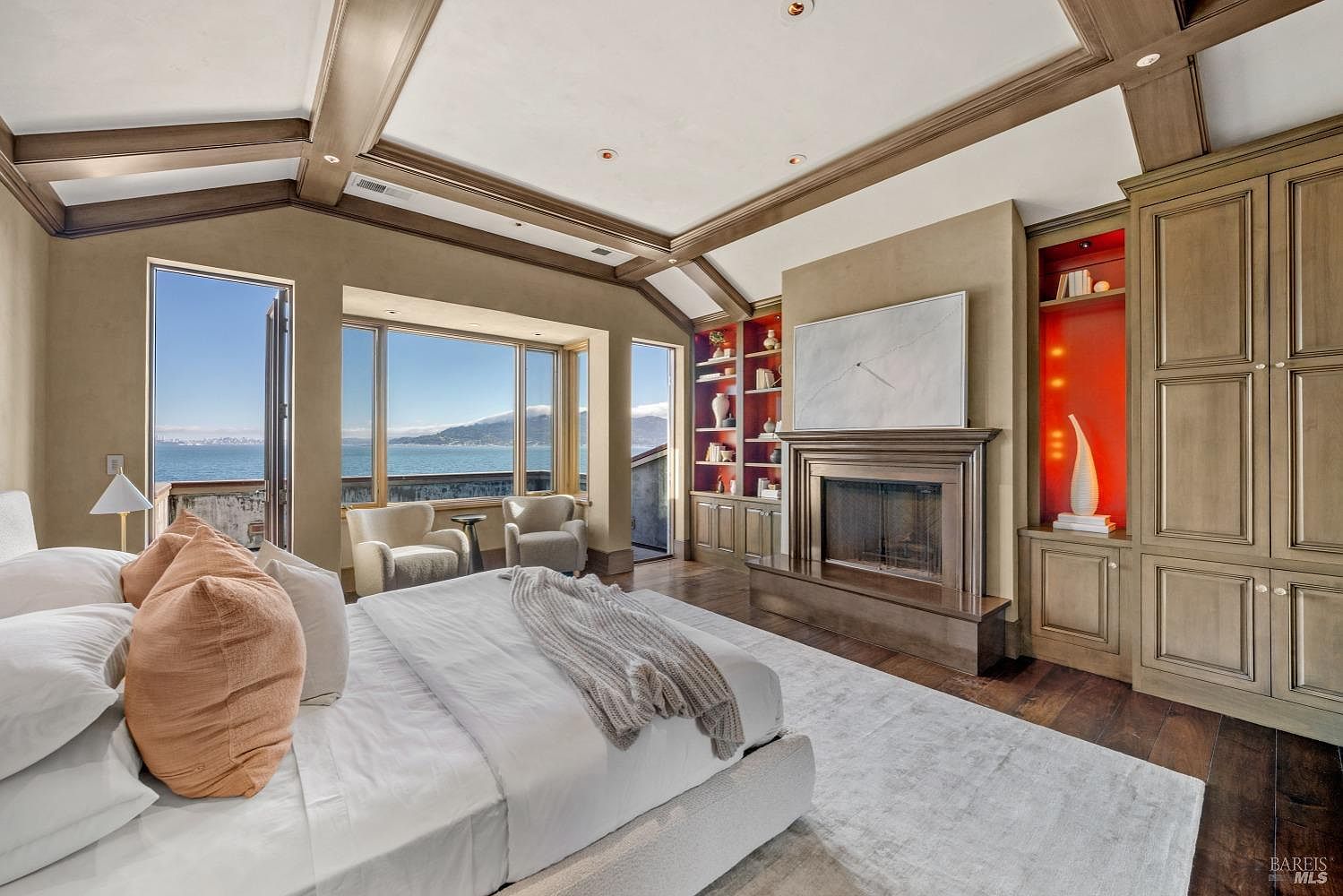 This is a luxurious primary bedroom featuring a king-size bed with white linens and decorative pillows, a fireplace with a marble surround and a large painting above, and custom built-in cabinetry with red-lit shelving. The room has a coffered ceiling with wood beams and large windows offering a stunning ocean view. The overall impression is one of elegance and tranquility.