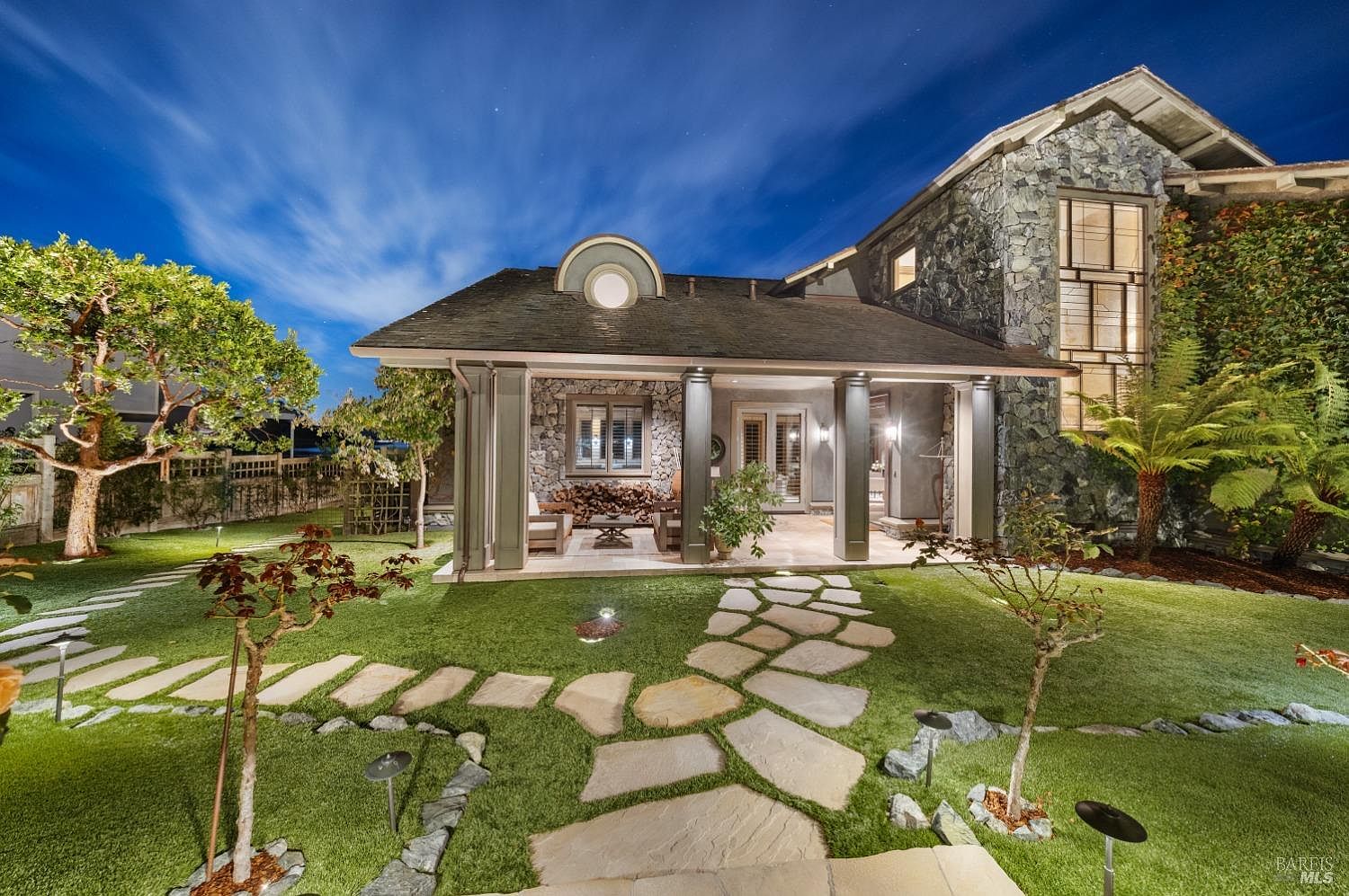 This image showcases the exterior yard and garden of a luxurious home at dusk. A stone pathway leads through a well-manicured lawn towards a covered patio area with stone pillars and a stone facade on the main house. The landscape is illuminated by subtle garden lighting, creating a warm and inviting ambiance.