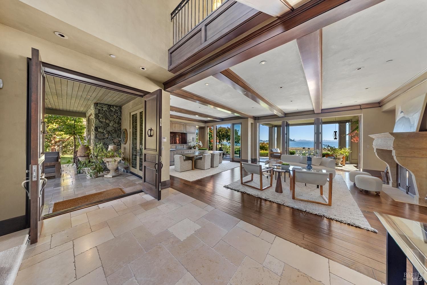 This is a spacious and luxurious living room that seamlessly blends indoor and outdoor living. The room features large windows with stunning water views, hardwood floors, and a neutral color palette. The open floor plan connects the living room to the dining area and kitchen, creating a cohesive and inviting space.