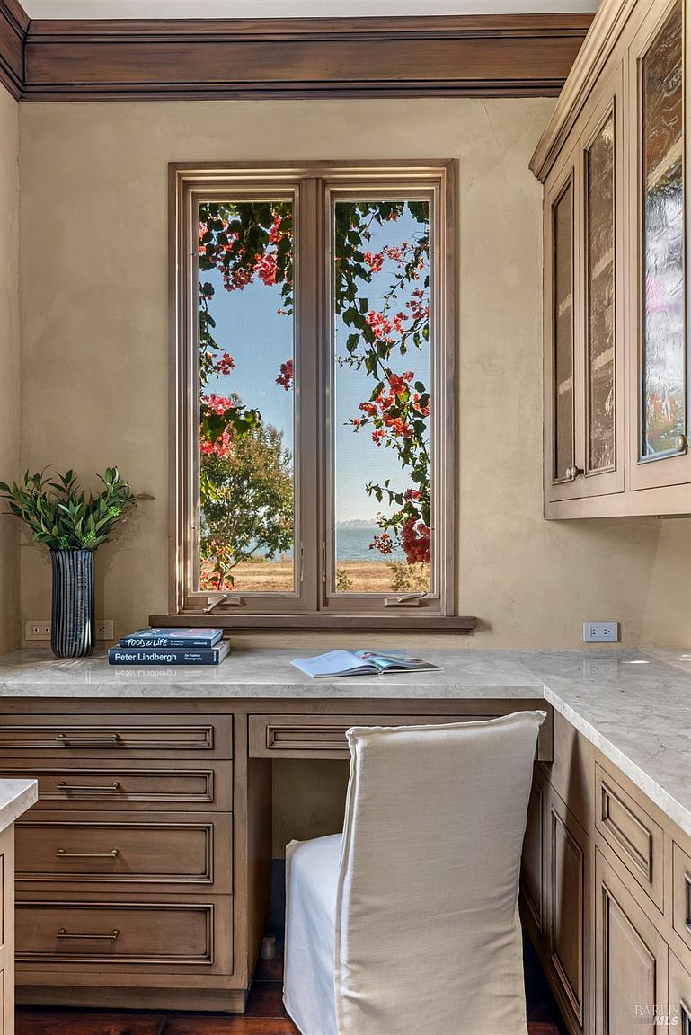 This interior shot showcases a cozy office or study area with custom cabinetry and a built-in desk. A window frames a picturesque view of the outdoors, adorned with flowering vines, adding a touch of nature to the workspace. The neutral color palette and natural light create a serene and inviting atmosphere, perfect for a home office.