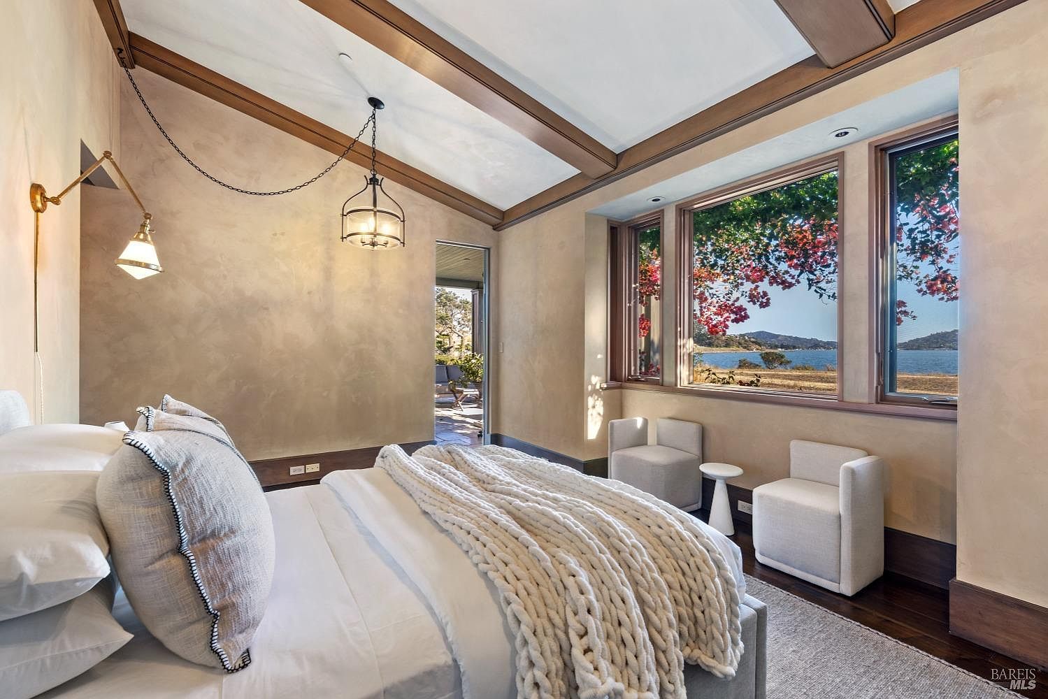 This is a primary bedroom featuring a king-size bed with white linens and a chunky knit throw. The room has a neutral color palette with textured walls and exposed wood beams on the ceiling. Large windows offer a view of the water, and there are two small chairs with a side table by the window. The perspective is from the foot of the bed, looking towards the windows and the doorway to the patio.