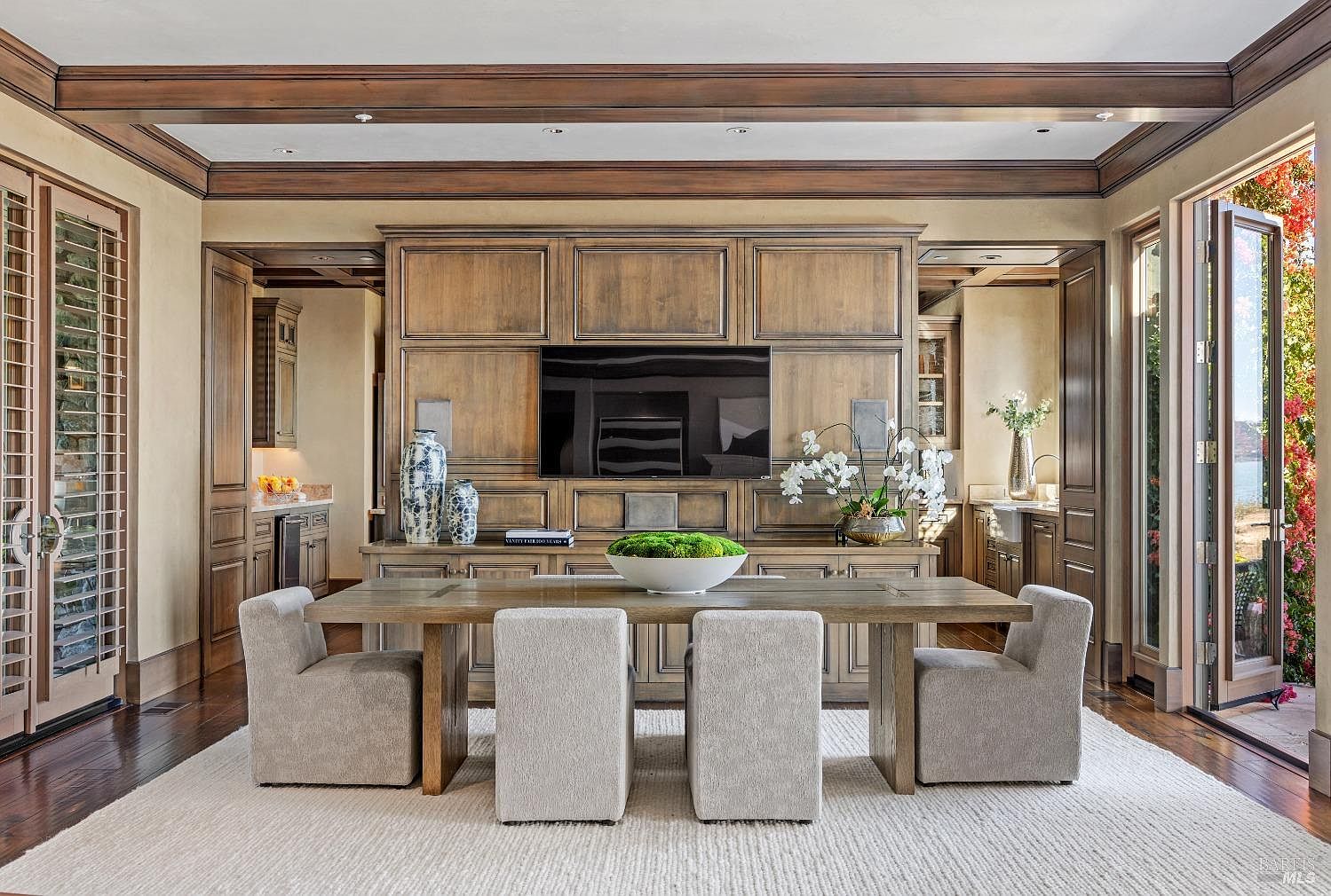 This is an interior shot of a dining room featuring a large wooden table with four upholstered chairs. A built-in wooden cabinet with a television above it serves as a focal point, flanked by doorways leading to other rooms. The room is well-lit, with natural light coming in from a set of folding doors on the right, and the overall style is traditional and luxurious.