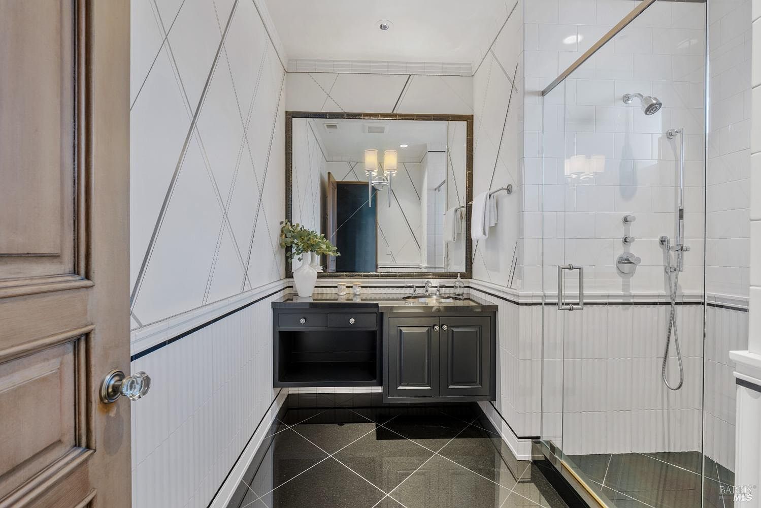 This is a stylish guest bathroom featuring a black vanity with a marble countertop and a vessel sink. The walls are decorated with a geometric pattern above a wainscoting detail, and the floor is covered in black marble tiles. A glass-enclosed shower with chrome fixtures is visible to the right, creating a modern and luxurious feel.