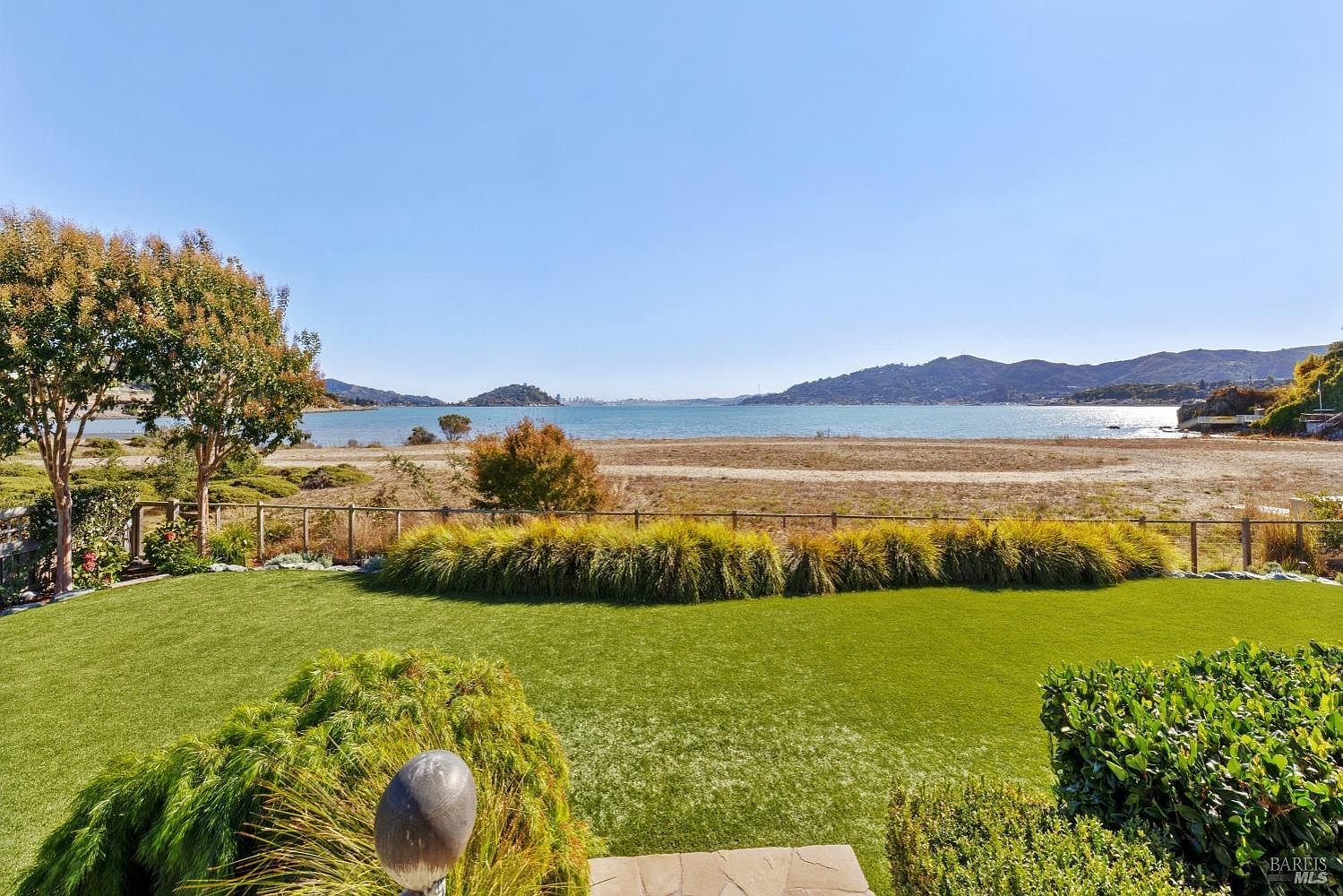 This image showcases a beautifully landscaped yard with a stunning waterfront view. The manicured lawn and well-maintained shrubs create an inviting outdoor space. A wooden fence separates the yard from the beach, offering a sense of privacy while still allowing for unobstructed views of the water and distant hills.