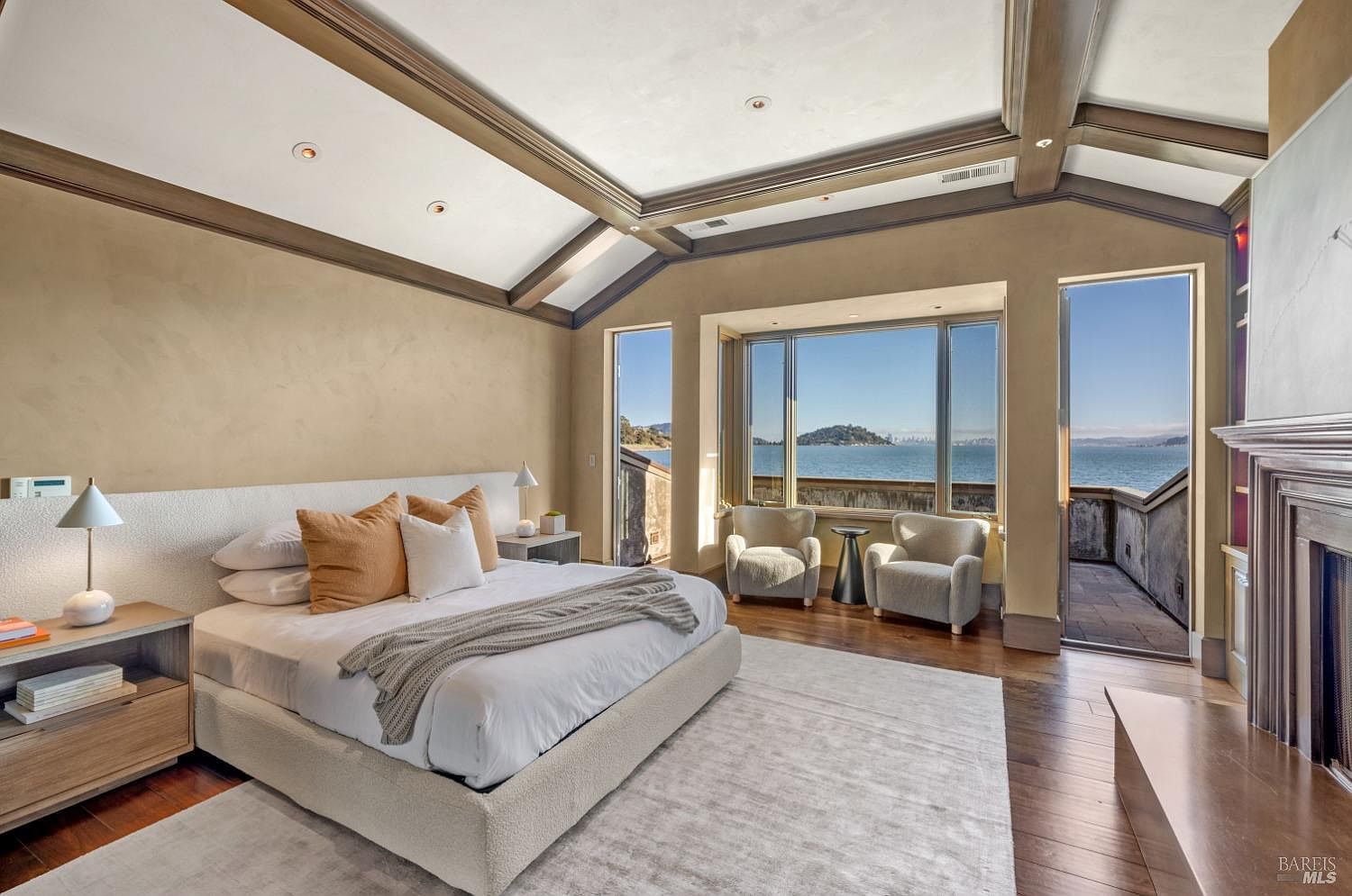This is a luxurious primary bedroom featuring a king-size bed with a textured headboard, complemented by neutral-toned bedding and decorative pillows. The room boasts a stunning ocean view through large windows, with two armchairs positioned to enjoy the scenery. The architectural details include exposed beams and a fireplace, adding to the room's elegance and appeal.