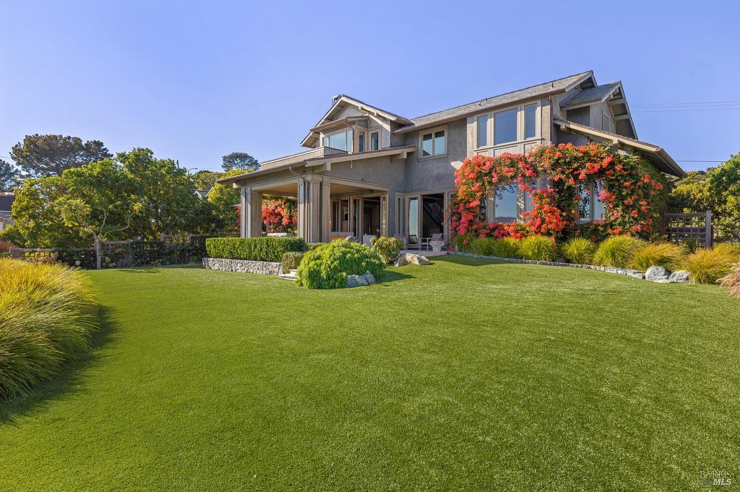 This image showcases the rear exterior of a well-maintained home with a lush green lawn. The house features a covered patio area supported by columns, large windows framed by vibrant orange flowering vines, and manicured landscaping. The overall impression is one of tranquility and sophisticated outdoor living.