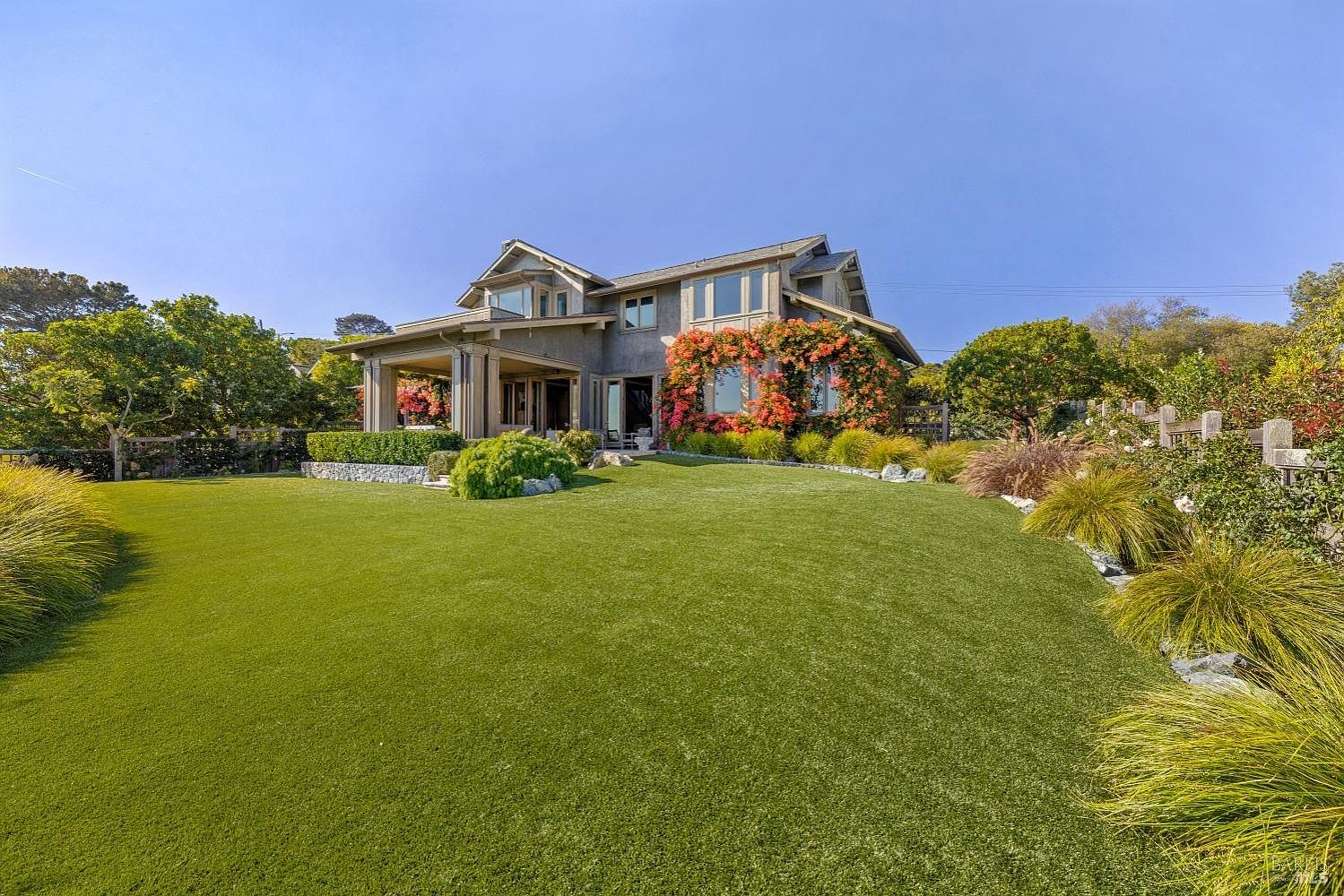 This image showcases a beautifully landscaped yard with a large, well-maintained lawn leading up to a substantial two-story home. The house features a neutral exterior with climbing flowering vines adding a pop of color, and the garden is thoughtfully designed with various plants, shrubs, and decorative rock borders. The overall impression is one of a serene and luxurious outdoor space.