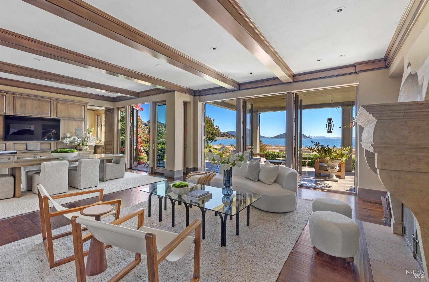 This is a luxurious living room featuring a coffered ceiling with wooden beams, hardwood floors, and a large area rug. The room opens to an outdoor patio with ocean views through folding glass doors, creating a seamless indoor-outdoor living experience. The furniture includes a curved sofa, glass coffee table, and modern chairs, contributing to an elegant and comfortable atmosphere.