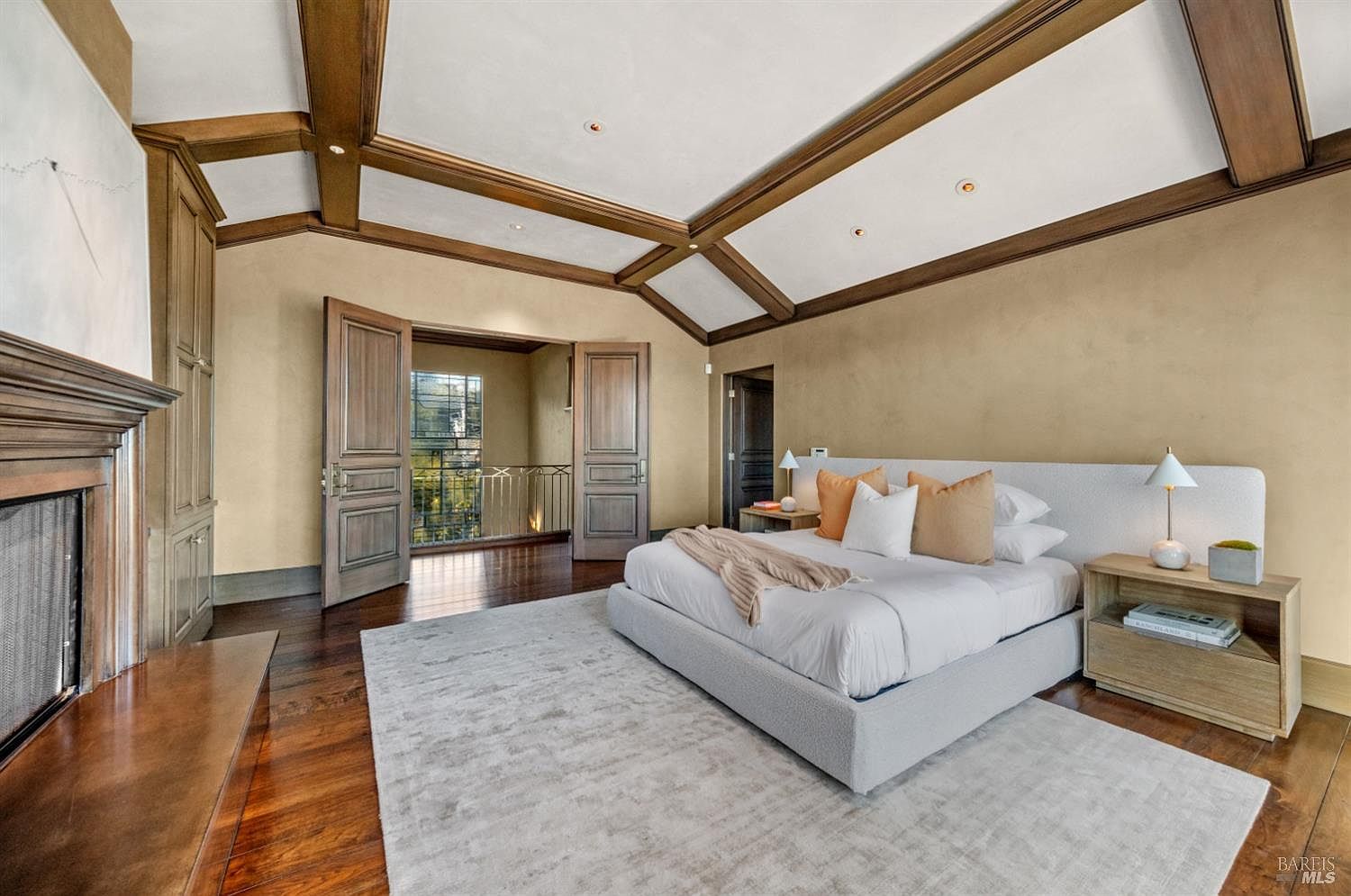 This is a primary bedroom featuring a large bed with a light gray upholstered frame, white linens, and decorative pillows. The room has hardwood floors, a light gray area rug, and a coffered ceiling with wooden beams. There is a fireplace on the left and doors leading to a balcony.
