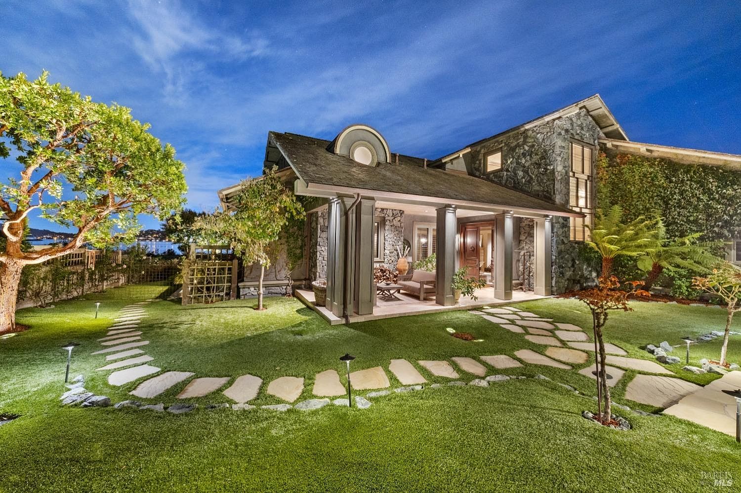 This image showcases a beautifully landscaped yard with a stone pathway leading to a covered porch of a stone house. The lush green lawn is accented by mature trees and decorative lighting, creating an inviting and serene outdoor space. The architecture of the house blends seamlessly with the natural surroundings, enhancing the property's curb appeal.