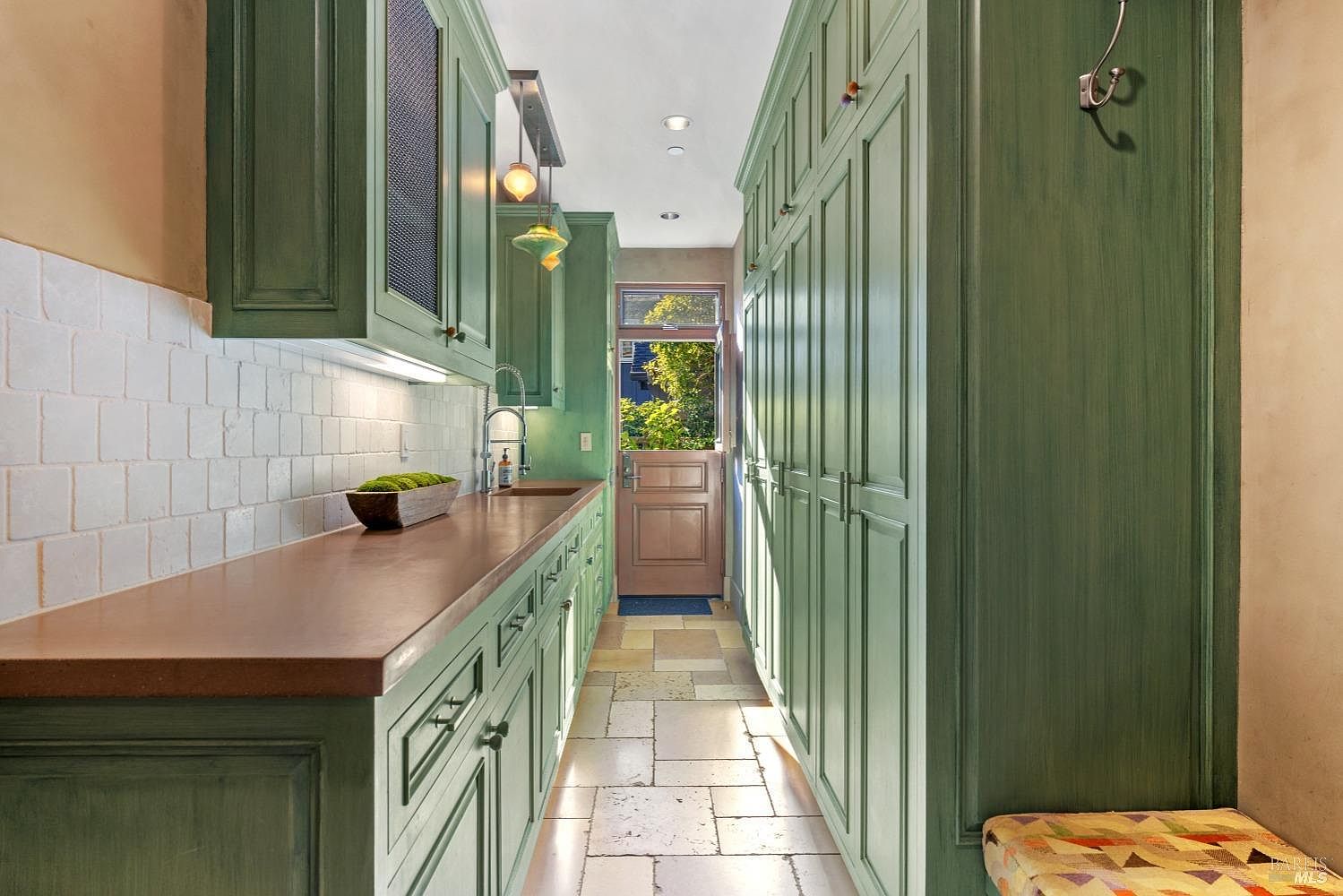 This interior shot showcases a well-organized laundry room with green cabinetry and a copper-colored countertop. The room features ample storage with floor-to-ceiling cabinets on one side and a sink area with overhead cabinets on the other. Natural light streams in through a door at the end of the room, highlighting the stone tile flooring.