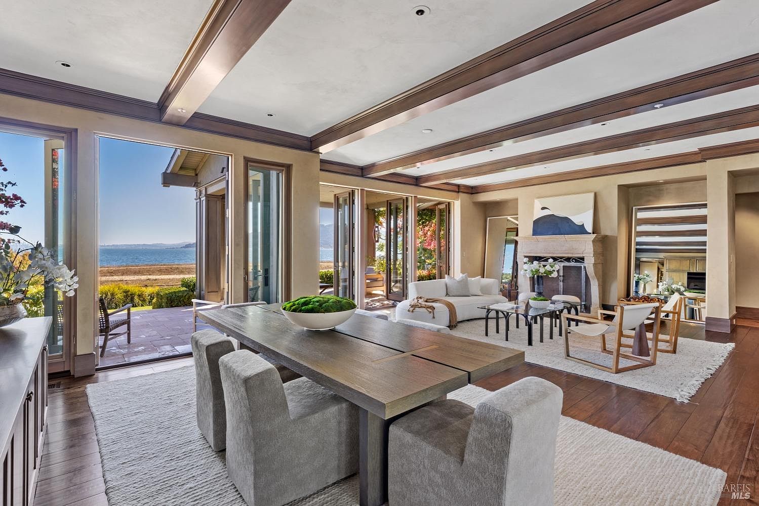 This is a luxurious living room featuring a dining area with a large wooden table and upholstered chairs, a cozy seating area with a white sofa and glass coffee tables, and a fireplace with an abstract painting above it. The room has hardwood floors, exposed ceiling beams, and large glass doors that open to an outdoor patio with ocean views, creating a seamless indoor-outdoor living experience. The perspective is wide-angle, showcasing the spaciousness and high-end finishes of the room.
