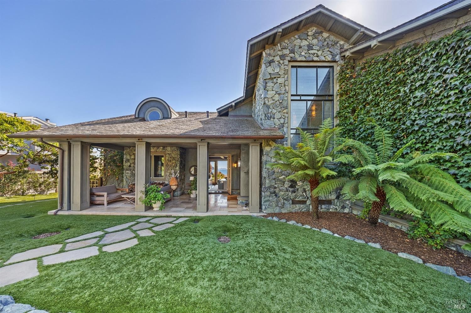 This is a front exterior view of a luxurious home featuring a stone facade, large windows, and lush landscaping. The entrance is framed by a covered porch with stone columns, leading to a well-manicured lawn and stone pathway. The overall impression is one of elegance and natural beauty.