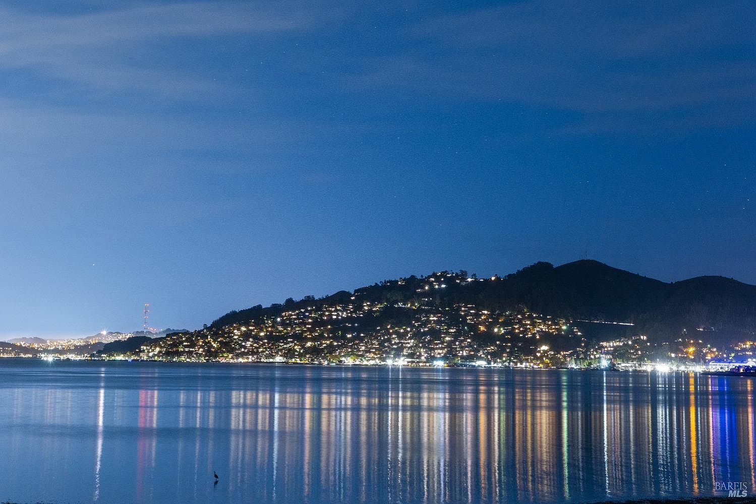 This nighttime aerial view showcases a hillside community with numerous illuminated homes reflecting in the calm water below. The lights create a warm and inviting atmosphere, highlighting the density and location of the residential area. The dark sky and water provide a dramatic contrast, emphasizing the community's presence.