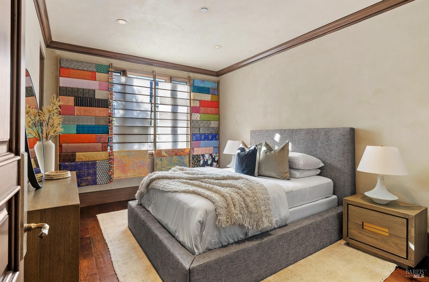 This is a well-lit primary bedroom featuring a gray upholstered bed with a plush throw blanket, two decorative pillows, and a nightstand with a modern lamp. The room has hardwood floors, neutral-toned walls, and a unique window treatment with colorful fabric panels. The overall impression is cozy and stylish.