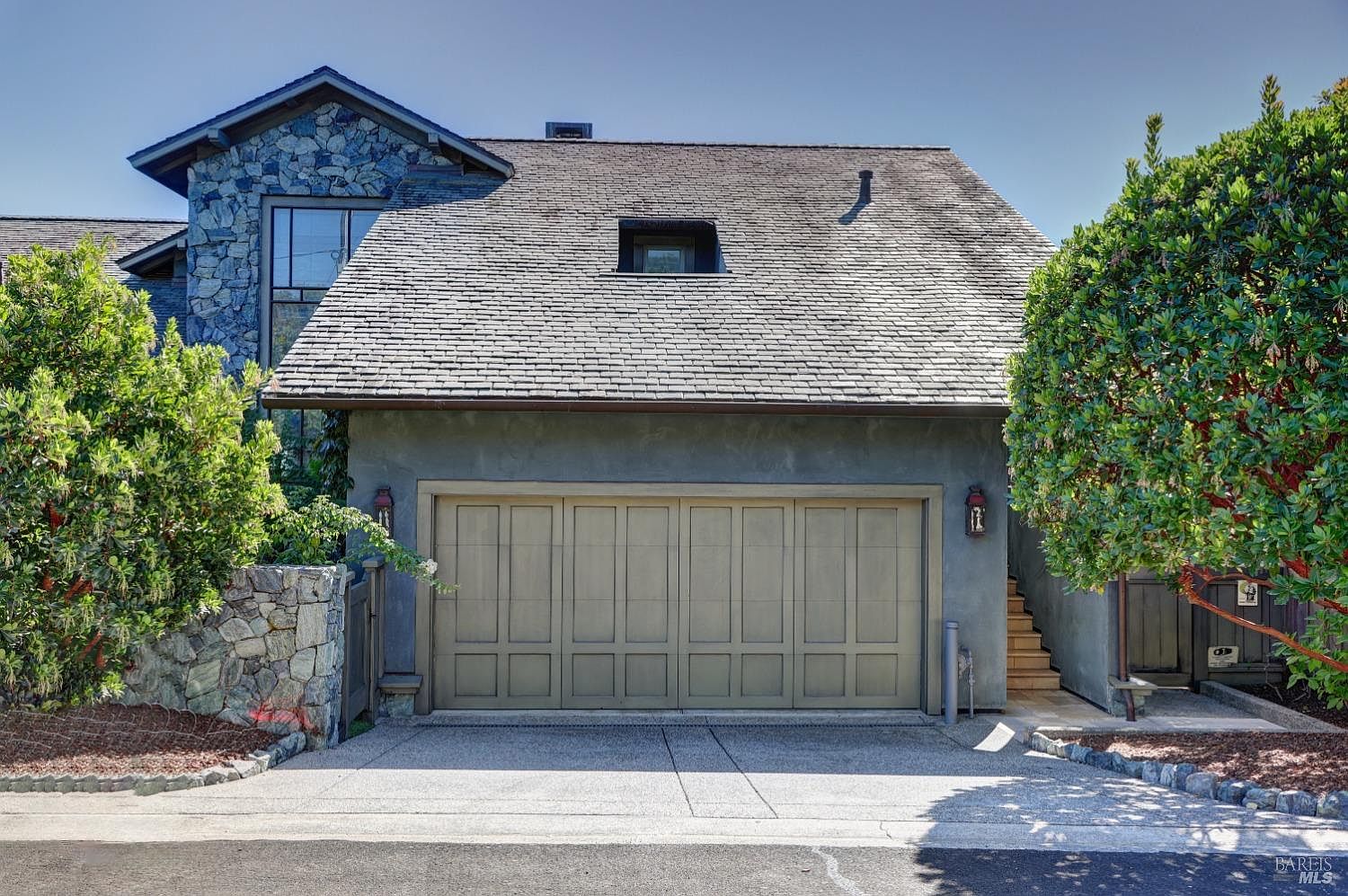This image showcases the exterior of a home, focusing on the garage. The garage features a multi-panel door, flanked by sconce lighting, and is set against a backdrop of a stone-accented wall and lush greenery. The driveway leads directly to the garage, presenting a clean and well-maintained facade.