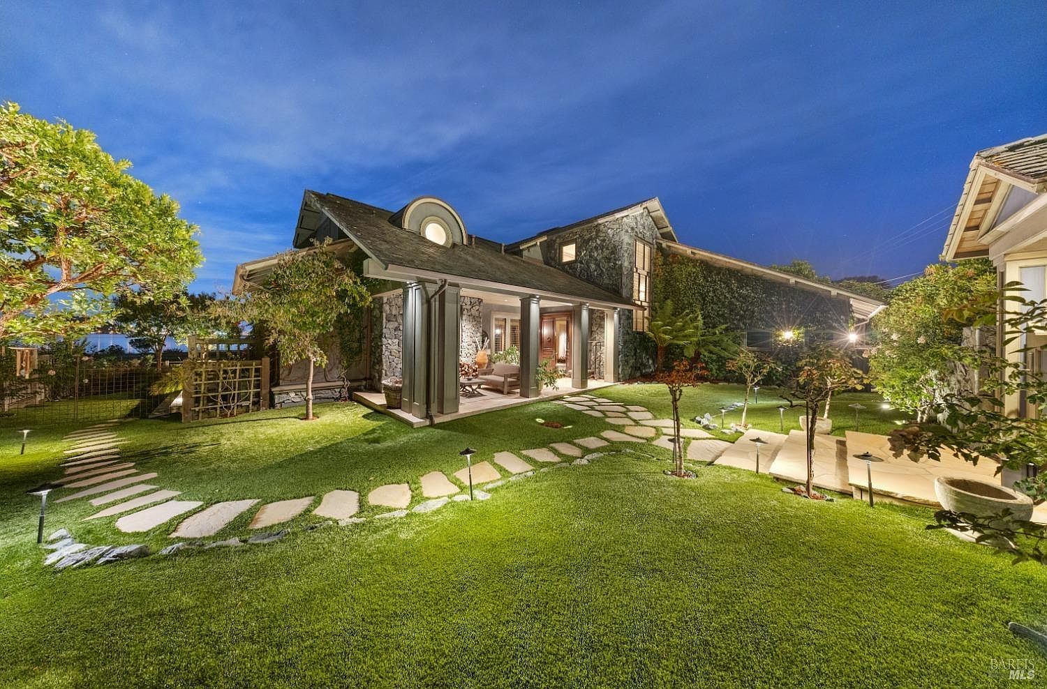 This image showcases a beautifully landscaped yard and garden at dusk, featuring a stone pathway leading to a stone-clad house with a covered porch. The lush green lawn is illuminated by subtle landscape lighting, creating a warm and inviting ambiance. The house is partially covered in ivy, adding to the natural aesthetic.