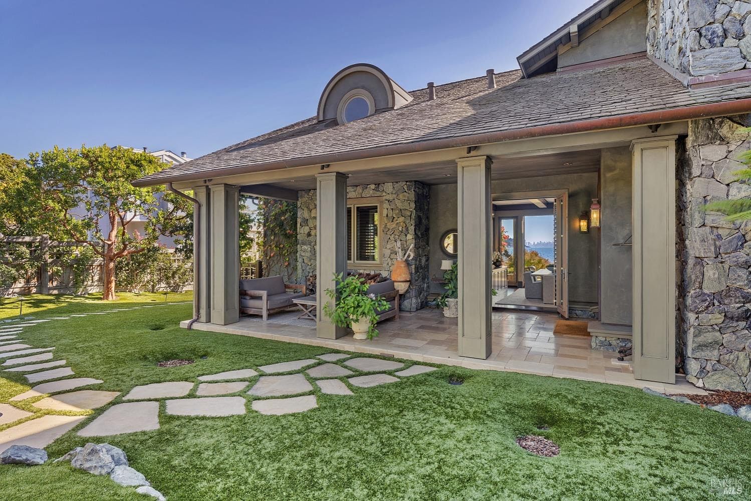 This image showcases the rear exterior of a luxurious home, featuring a covered patio with stone columns and comfortable seating. A well-manicured lawn with stone pathways leads to the patio, creating an inviting outdoor space. The architecture incorporates stone and wood elements, lending a sophisticated and natural aesthetic.